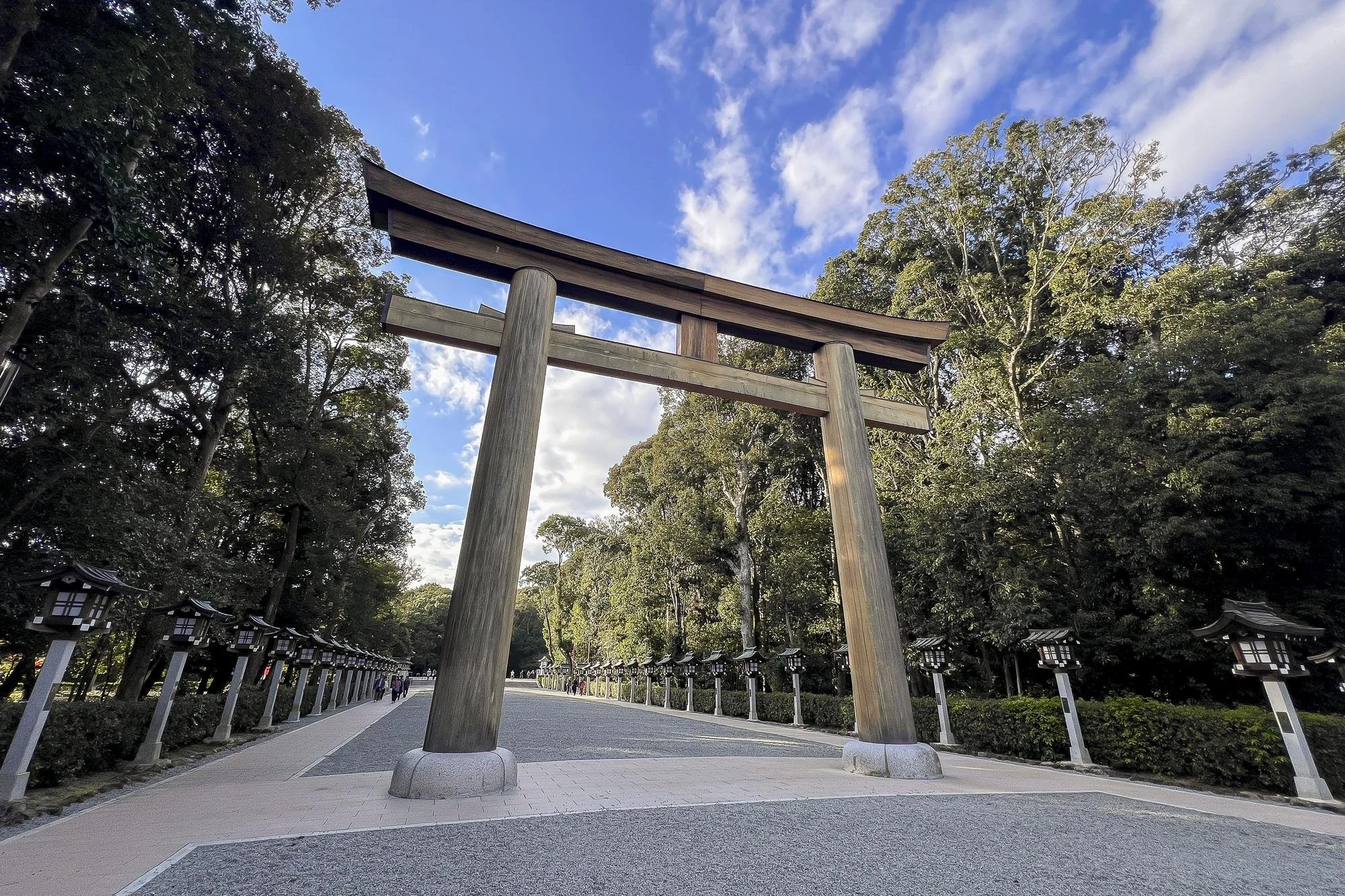A traditional Japanese torii gate made of wood, situated in a park with trees and lanterns along the pathway under a partly cloudy sky.