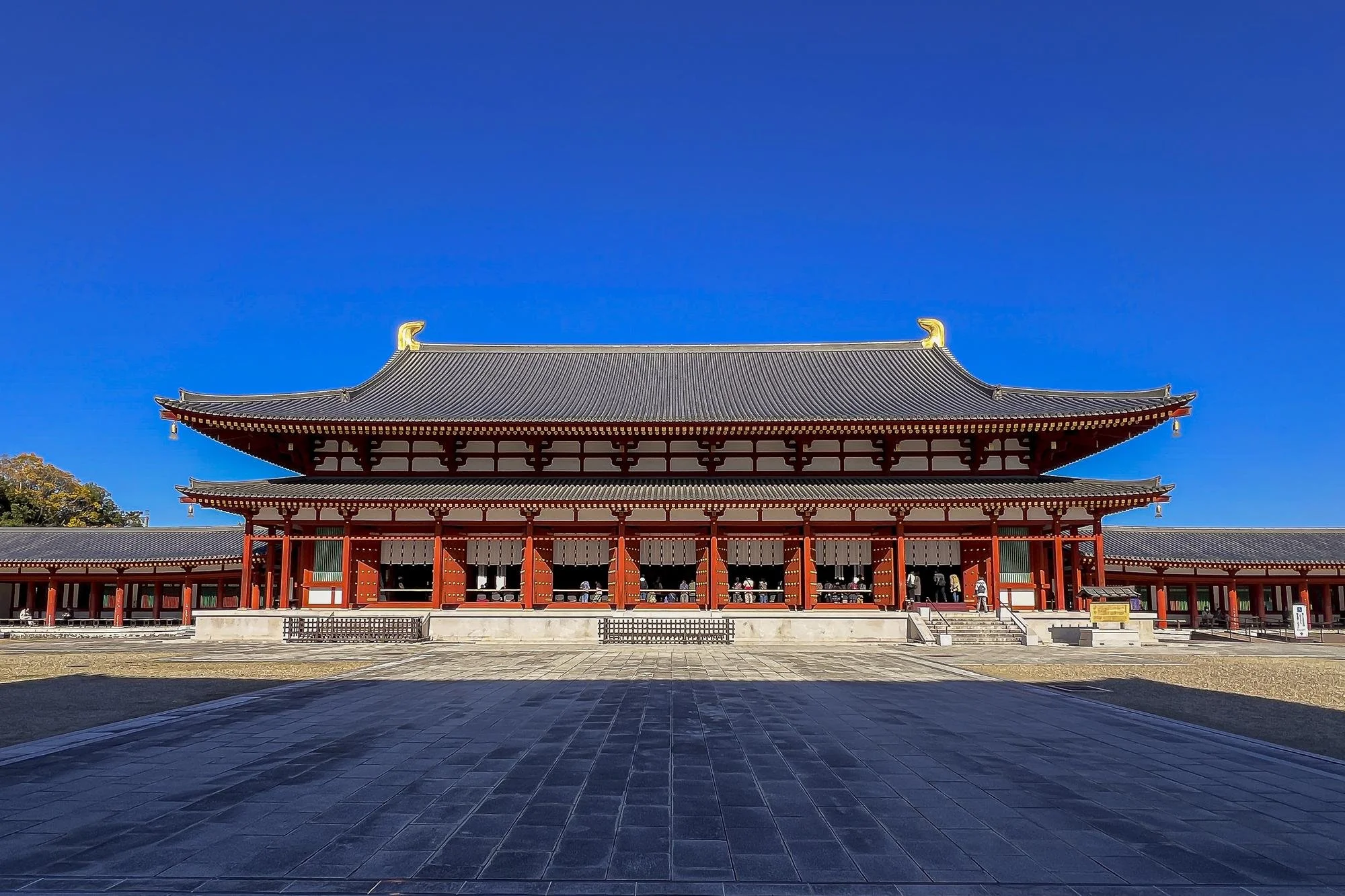 Traditional Japanese temple with red wooden beams and dark gray tiled roof against a clear blue sky.