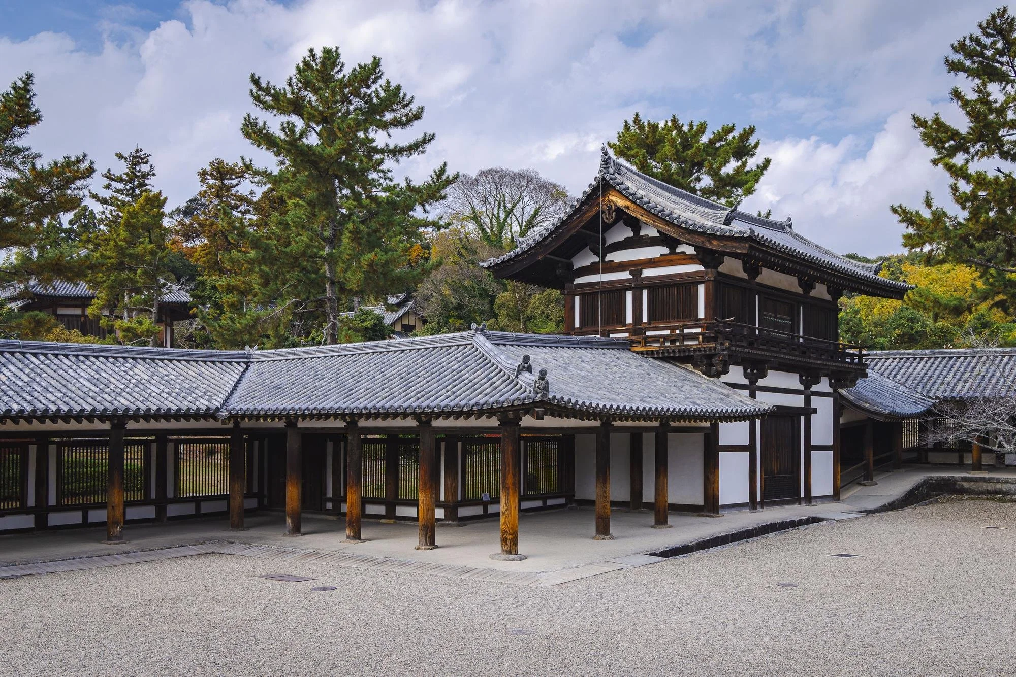 Traditional Japanese wooden building with tiled roof, surrounded by trees and a sandy courtyard.