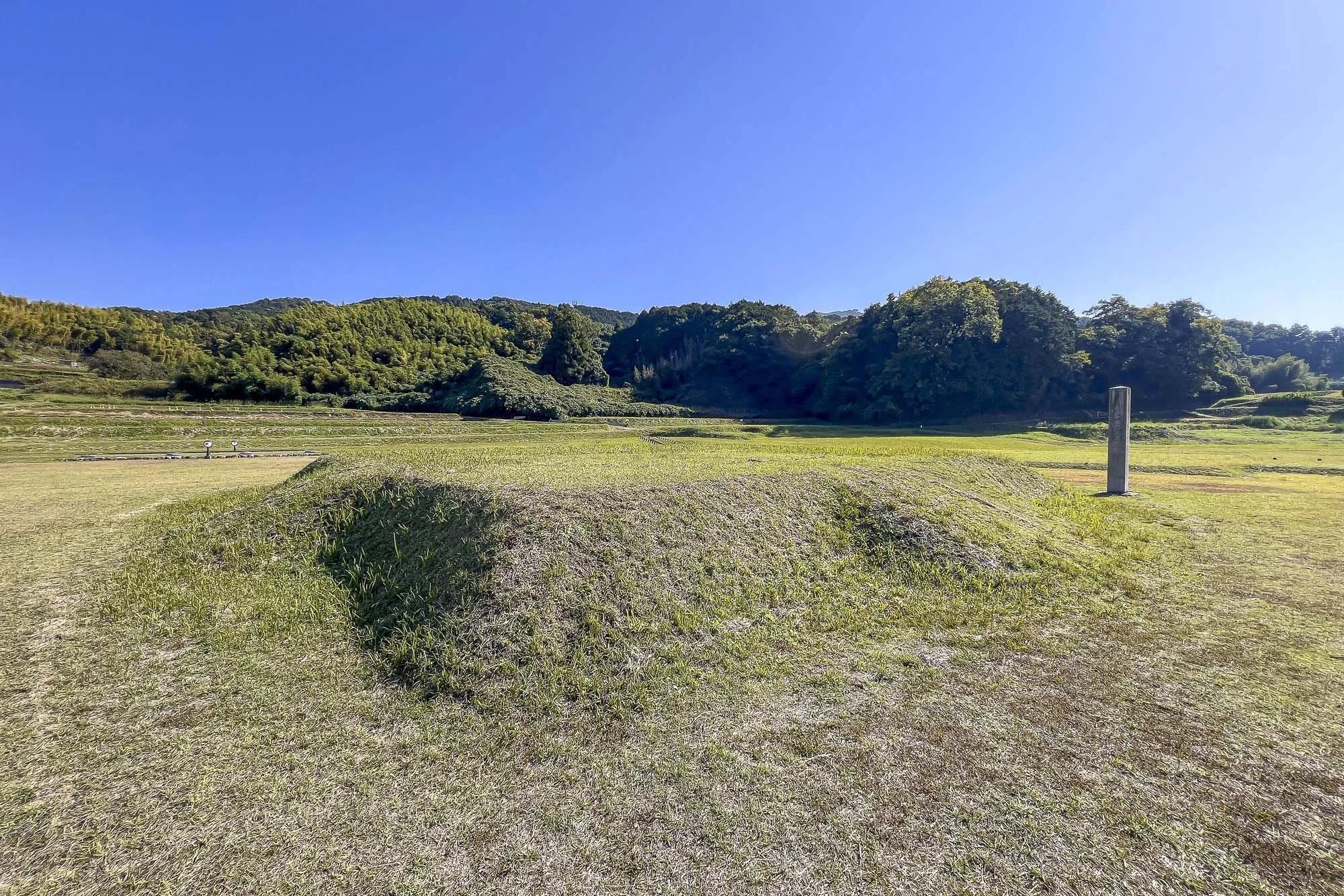 Open grassy landscape with a mound and a tall stone pillar, surrounded by hills and trees under a clear blue sky.