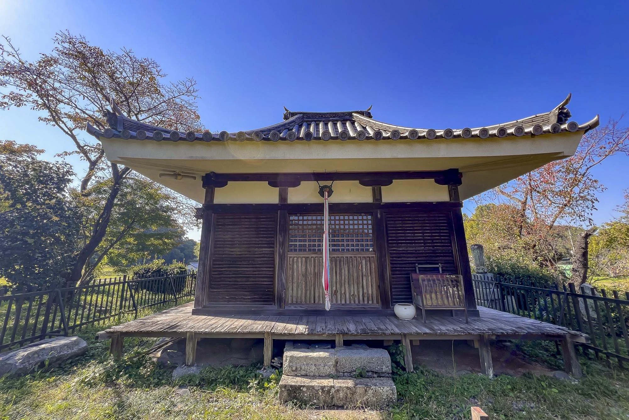 A traditional Japanese wooden shrine with a curved roof, wooden steps leading up to the entrance, surrounded by trees with autumn foliage, and a blue sky overhead.