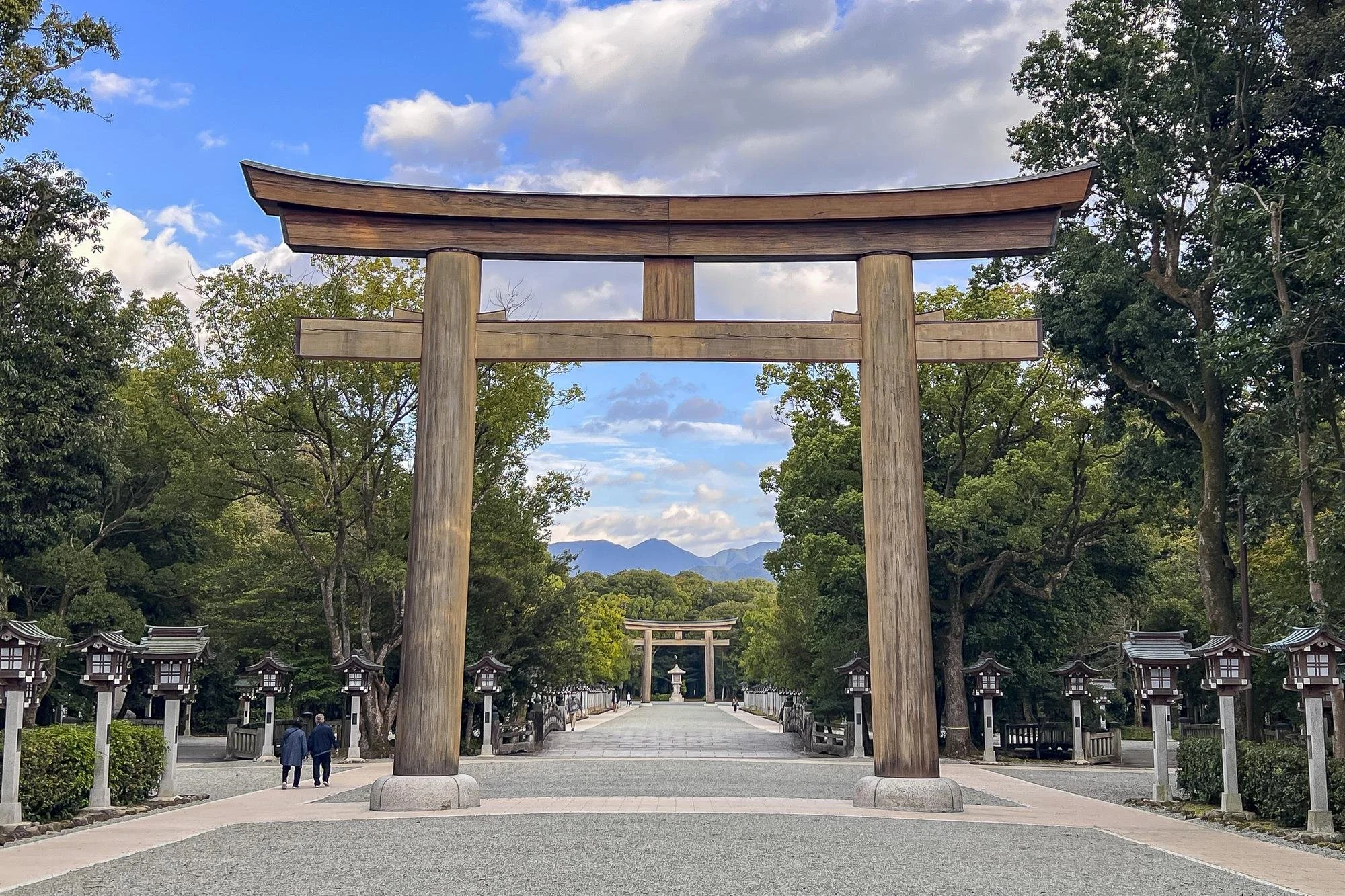 Large wooden torii gate leading to a traditional Japanese shrine path with trees and smaller torii gates in the background, mountain mountains in the distance, and a partly cloudy sky.