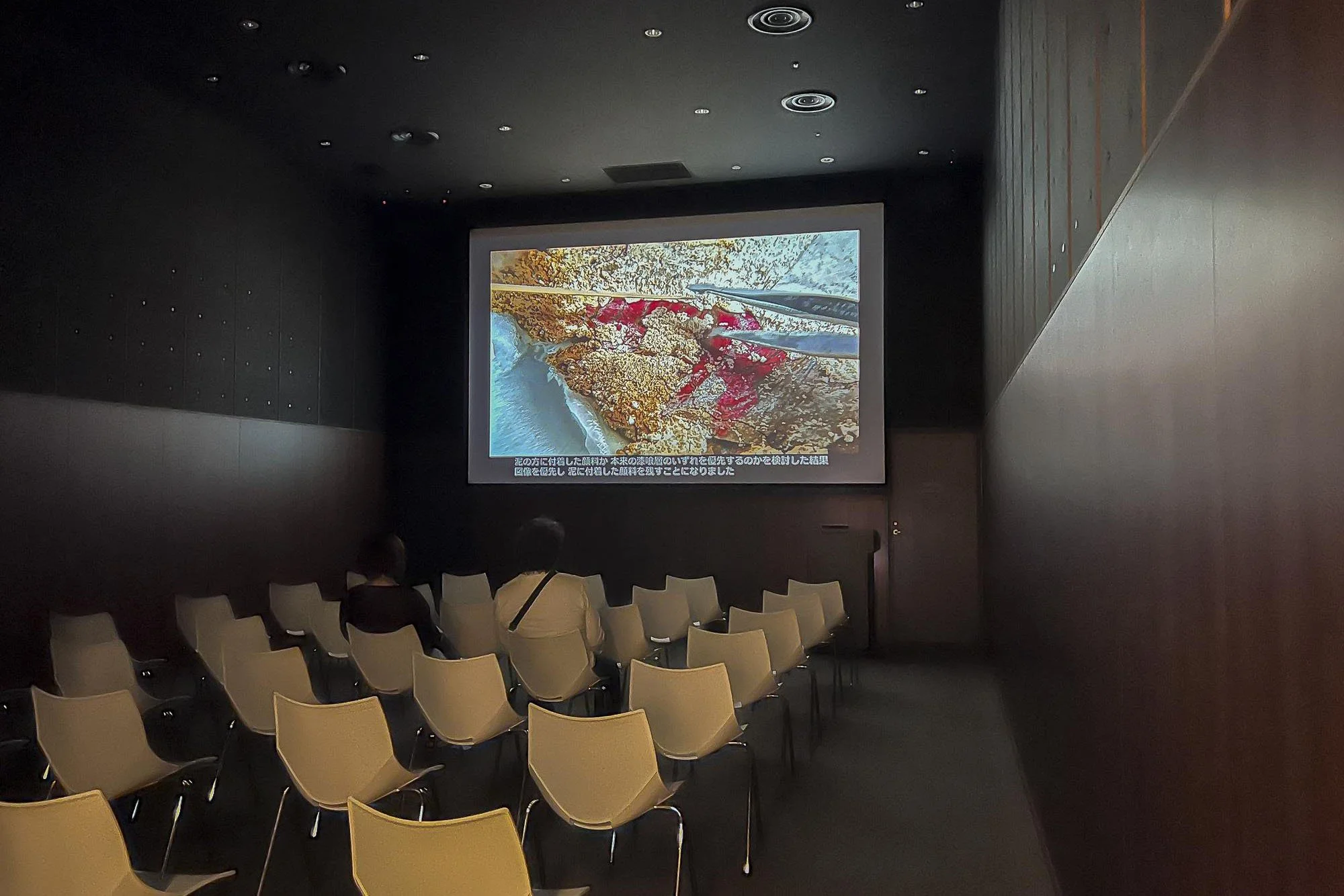 Two people sitting in chairs watching a presentation with a large screen displaying a nature scene and Japanese subtitles.