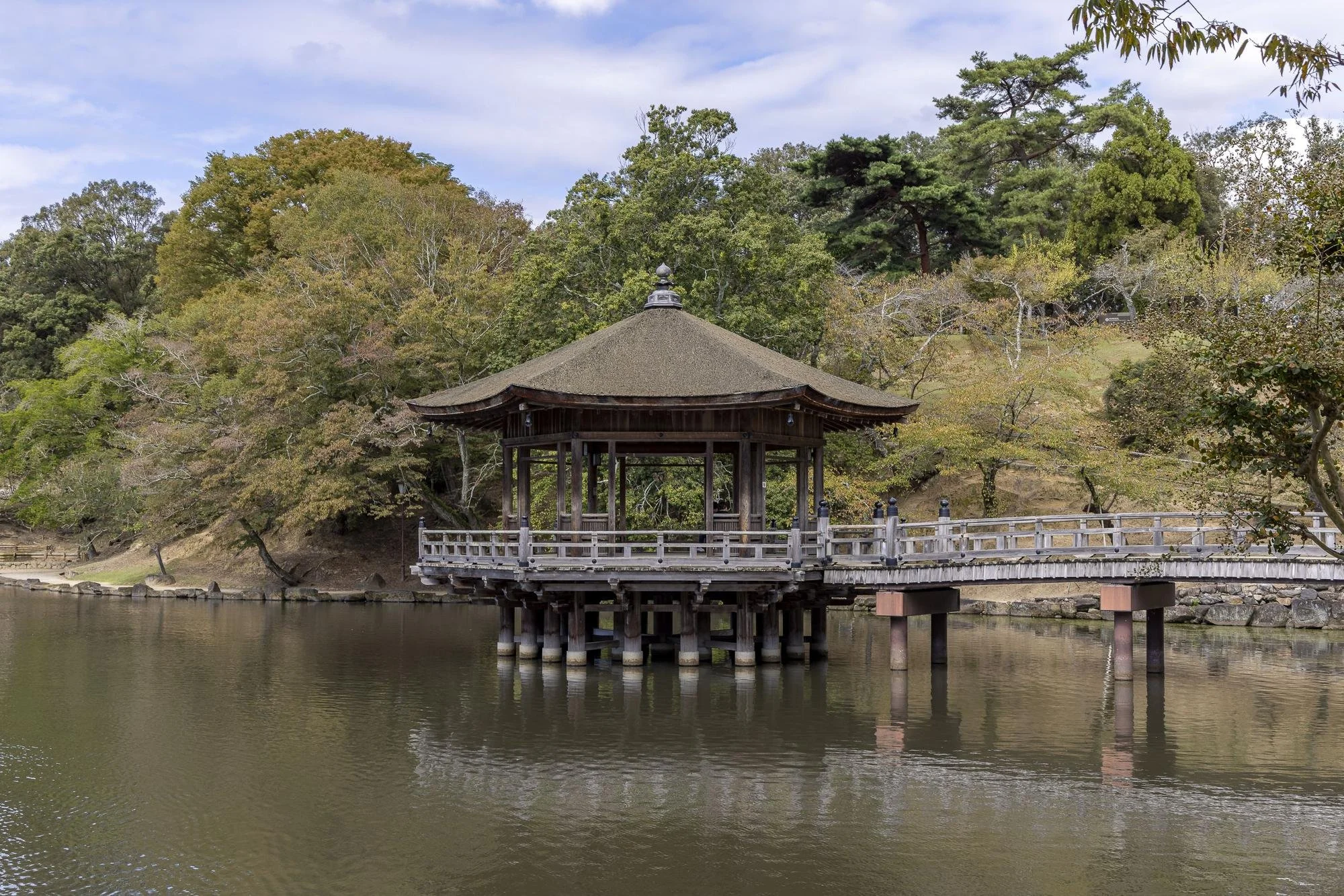 A traditional Japanese wooden gazebo on stilts over a calm body of water, with trees and a cloudy sky in the background.