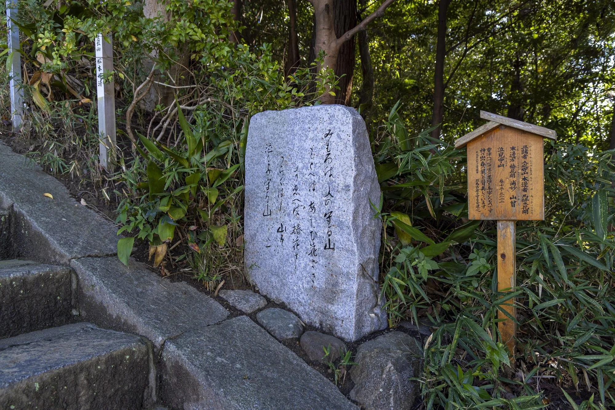 A large upright gray stone with Japanese calligraphy on it, situated among greenery and stone steps. To the right of the stone, there is a small wooden plaque with Japanese writing, mounted on a wooden post. Trees and bushes surround the area, creati