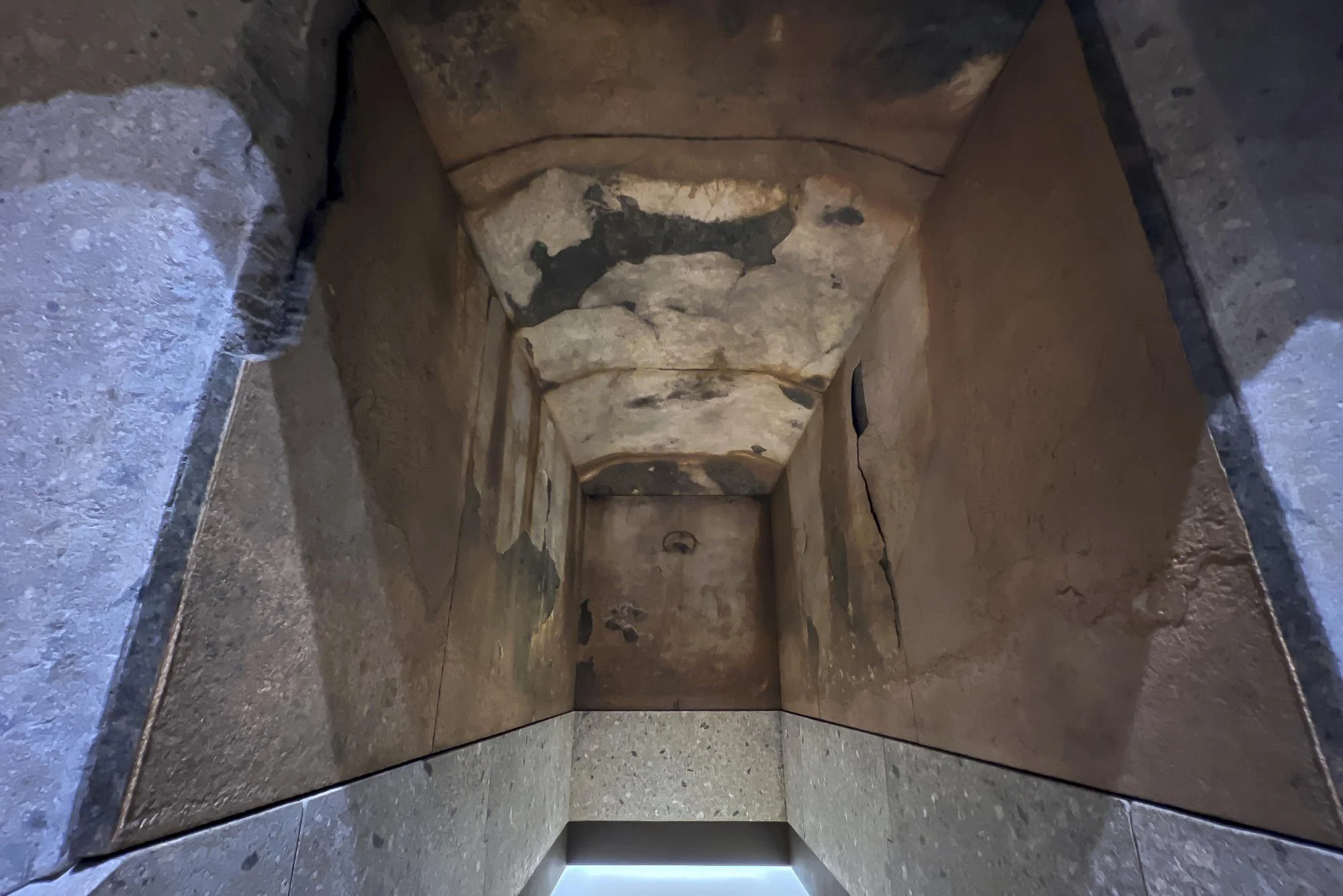 View of an old, narrow corridor with weathered stone walls and peeling plastered surfaces, leading to a small, dark doorway at the end.