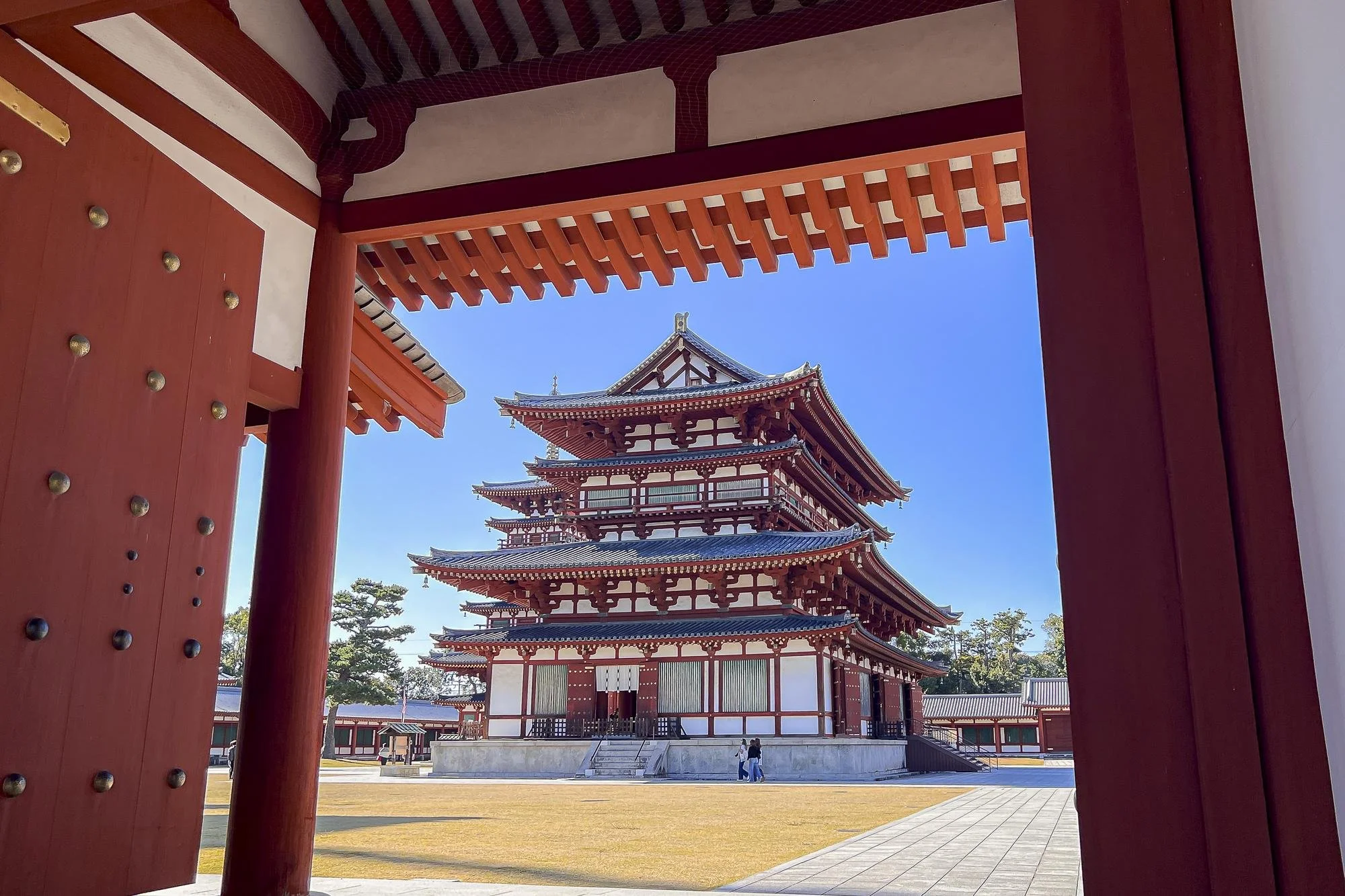 Traditional Japanese temple or pagoda seen through an open red wooden gate with a clear blue sky in the background.