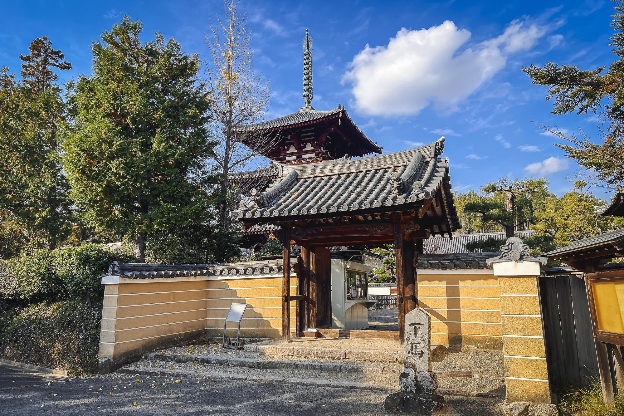 Traditional Japanese temple gate with tiled roof, wooden beams, and stone steps, surrounded by trees and a blue sky.