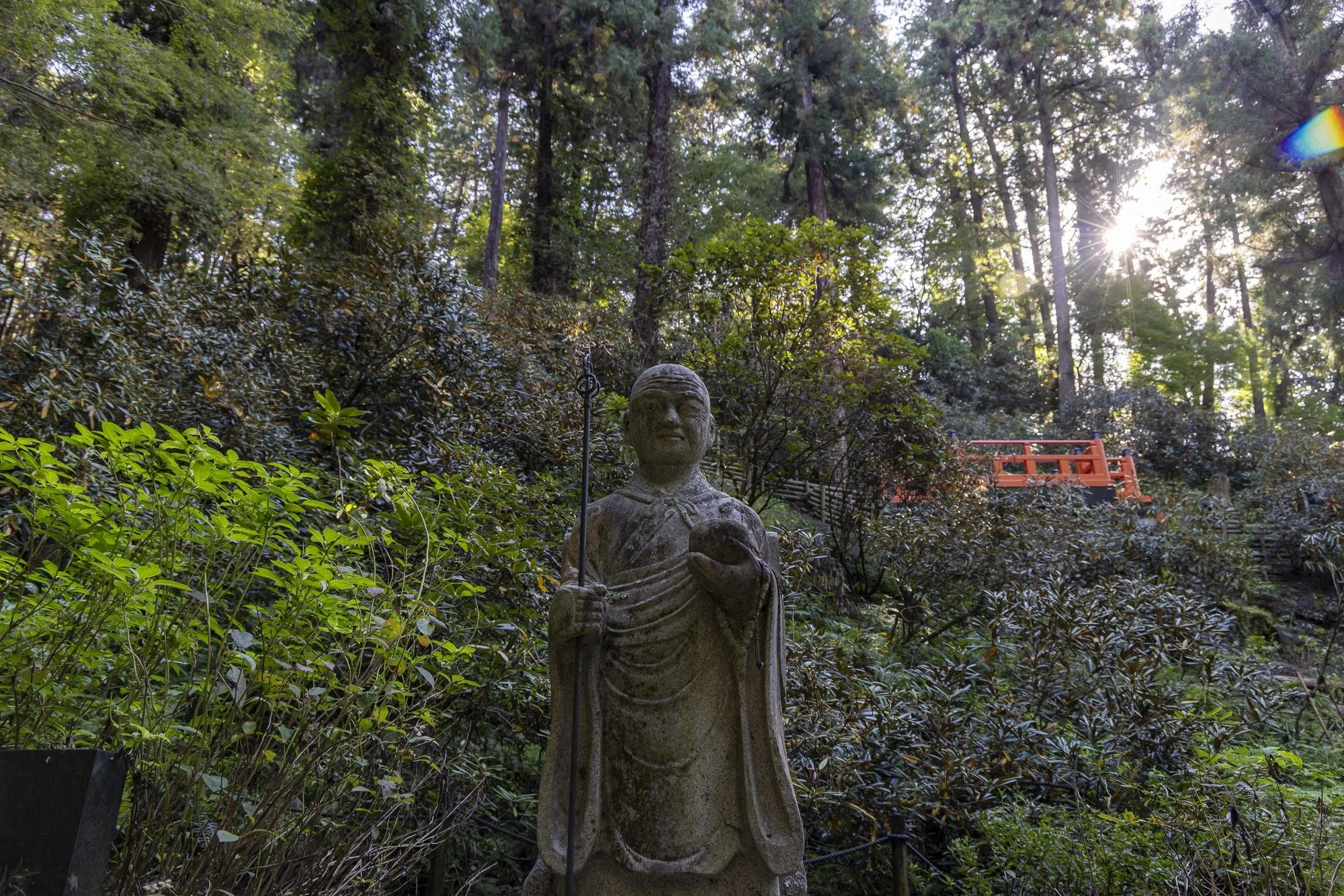 A stone statue of a person holding an object, standing among lush greenery with tall trees in the background. Sunlight filters through the trees, creating a bright spot near the top right.