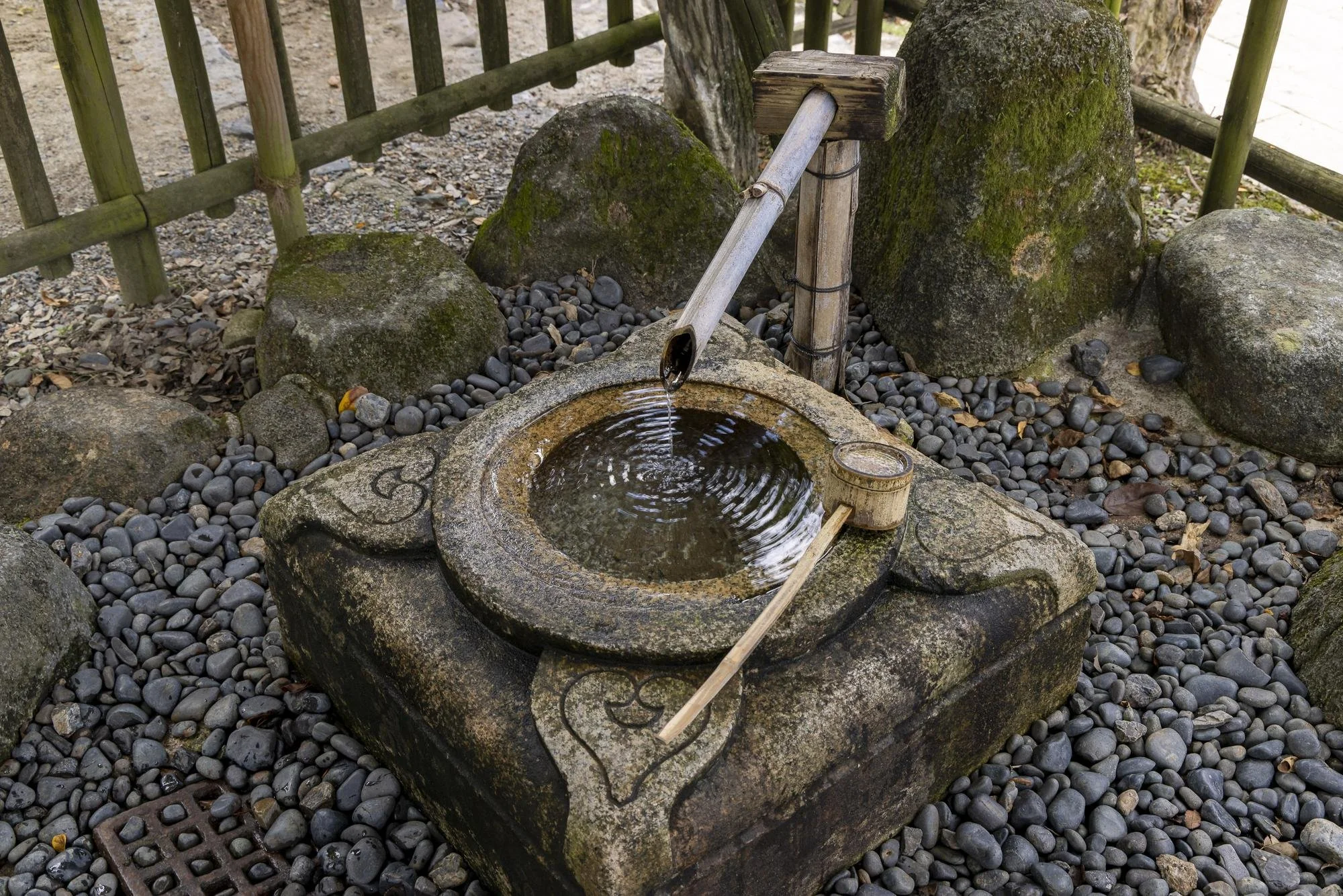 Traditional stone water basin with bamboo spout, used for purification at a Japanese shrine or temple, surrounded by rocks and wooden fence.