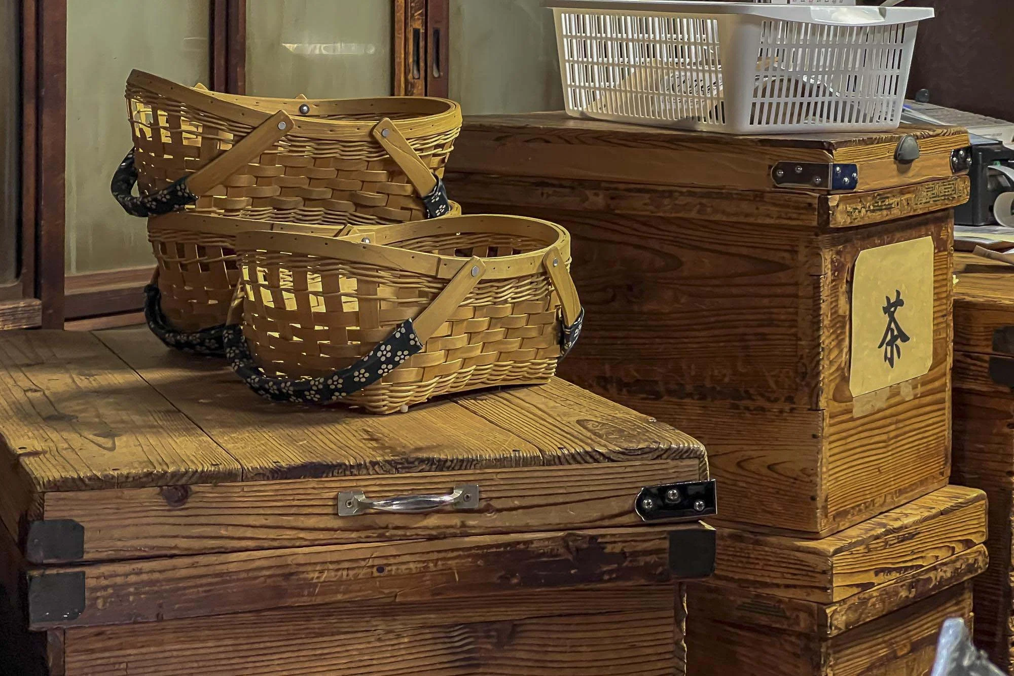 Two stacked wicker baskets with black fabric handles and a wooden box with a Japanese character label, on a wooden surface.