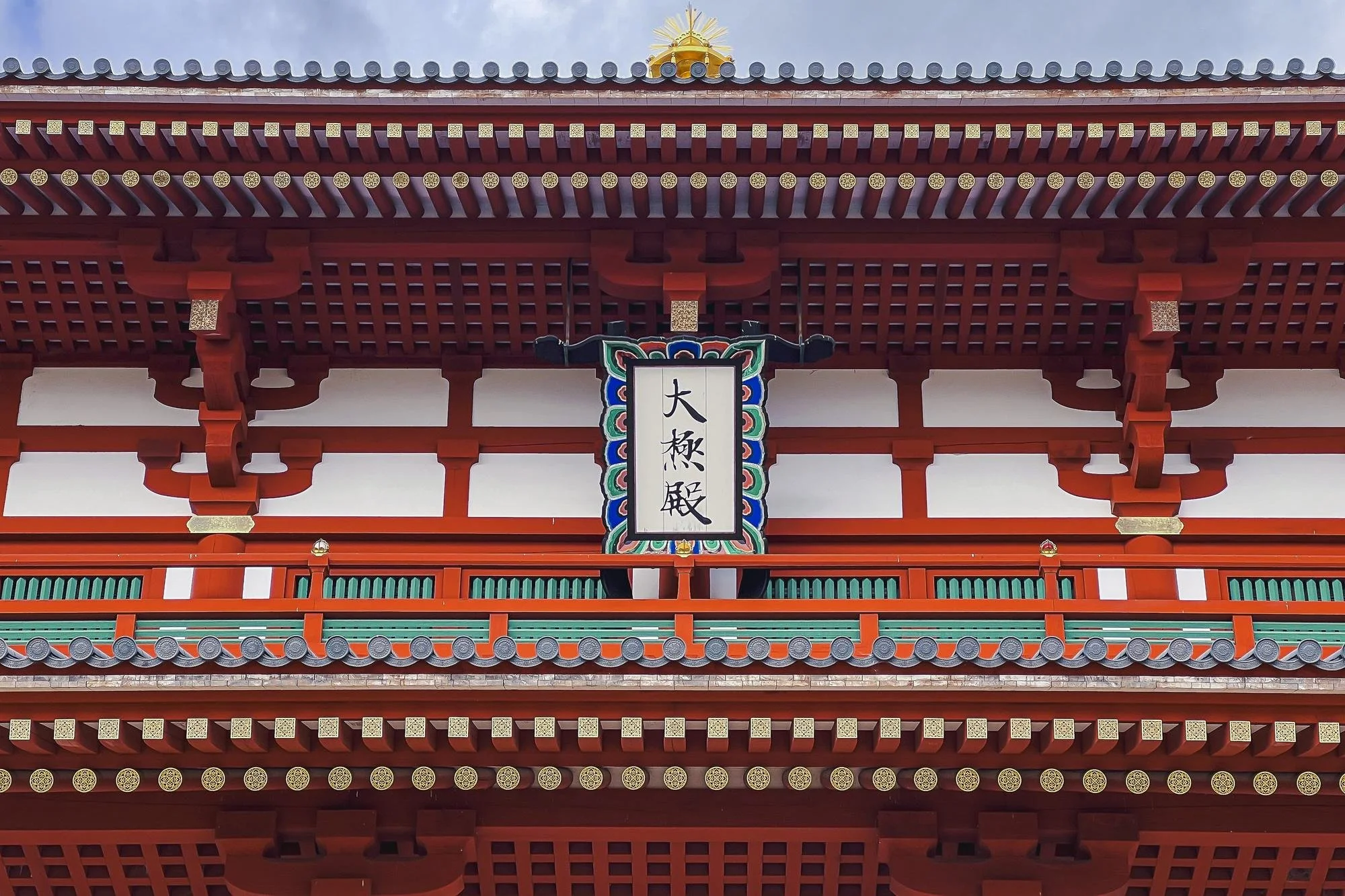 Close-up view of a traditional Japanese temple's upper balcony, featuring red wooden beams, ornate detailing, and a hanging sign with Japanese writing, set against a blue sky background.