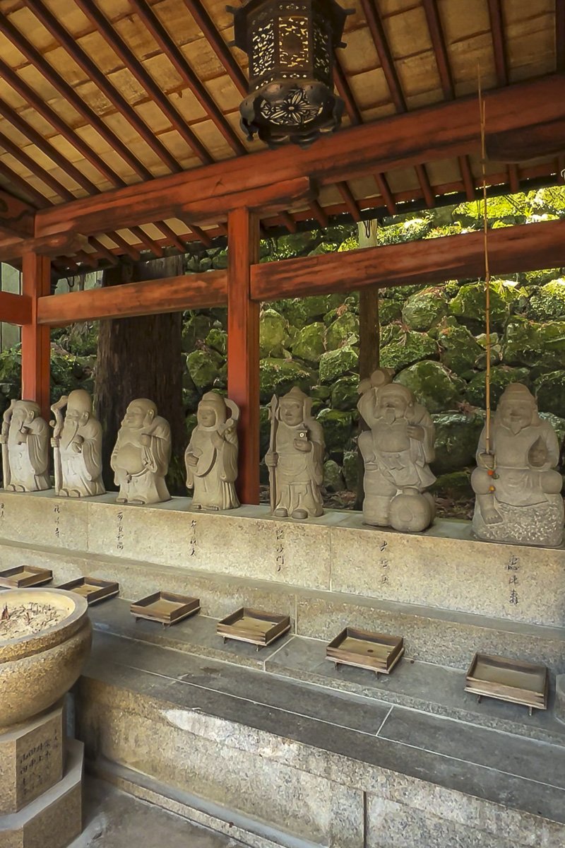 Series of small stone statues of Buddhist monks or deities seated and standing, arranged on a stone ledge in a traditional Japanese setting with a wooden roof and green foliage in the background.