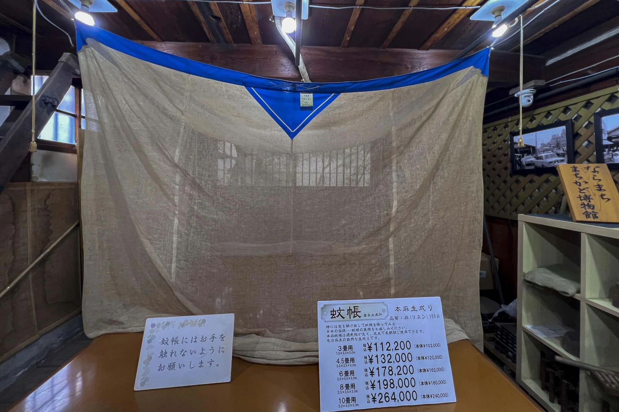 A beige mosquito net with a blue top hanging from the ceiling in a store display. There are price tags and informational signs in Japanese in front of it, along with a small shelf and framed pictures on the wall.