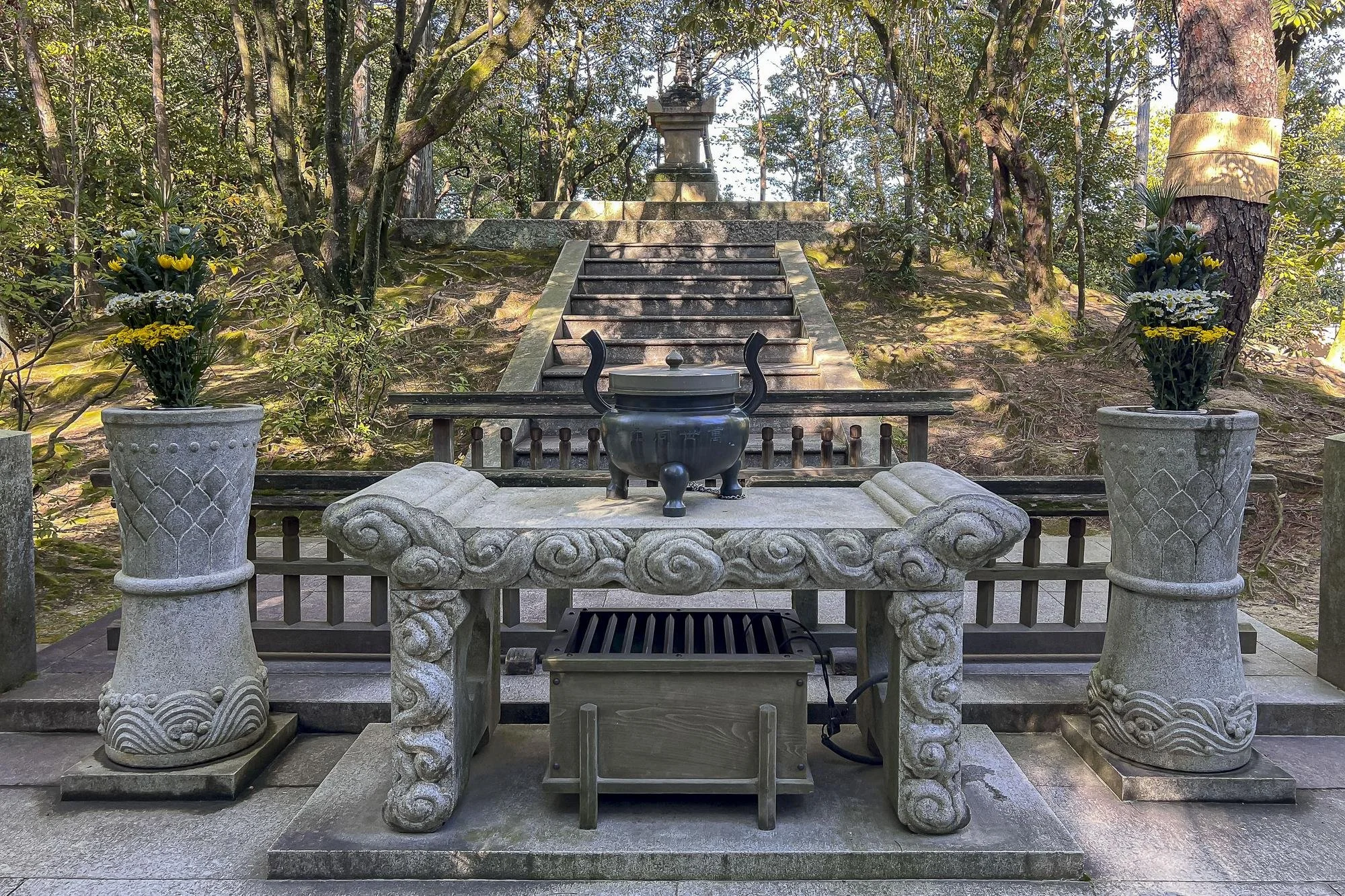 Stone altar with a black incense vessel, flanked by two tall stone vases with yellow and white flowers, set in a forested area with stairs leading up to a statue on a hill.