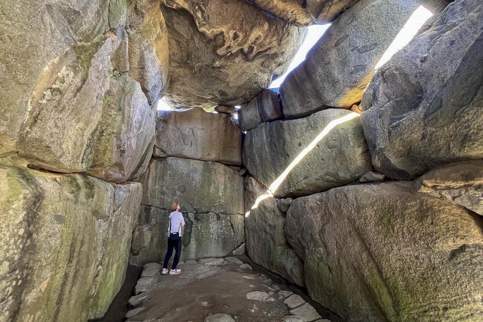 Person standing inside a large narrow rock formation with big stones and cracks, sunlight streaming through openings above.