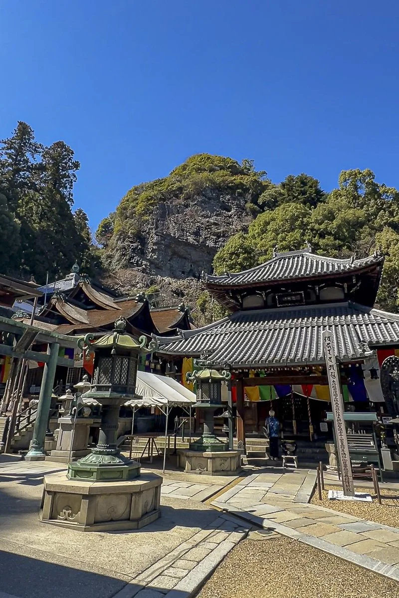 A traditional Japanese temple with ornate rooftops, stone lanterns, and colorful banners, set against a backdrop of green trees and a rocky hillside under a clear blue sky.