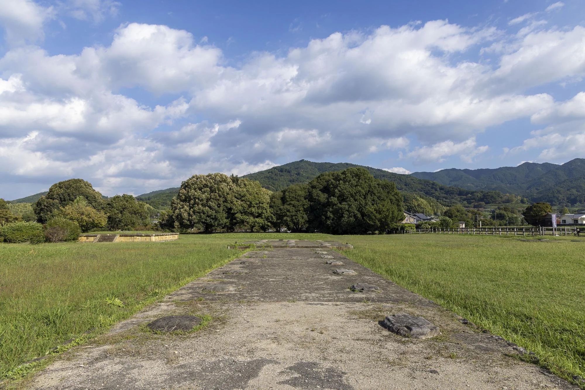 A paved pathway in a grassy field leads towards green trees and mountains in the distance under a partly cloudy sky.
