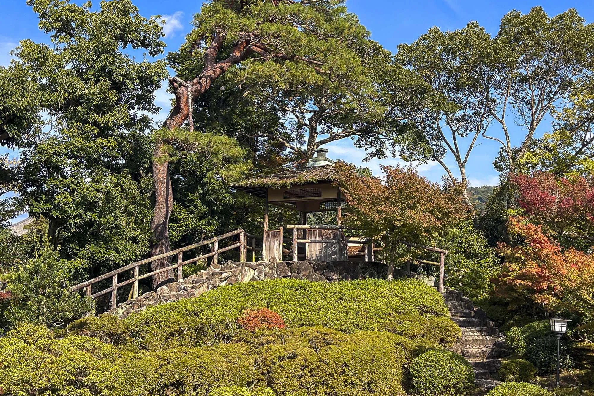 A small wooden pavilion with a tiled roof is situated on a hill surrounded by lush greenery, colorful trees, and a stone staircase leading up to it. There is a classic street lamp on the right side of the image, and the sky is partly cloudy with blue