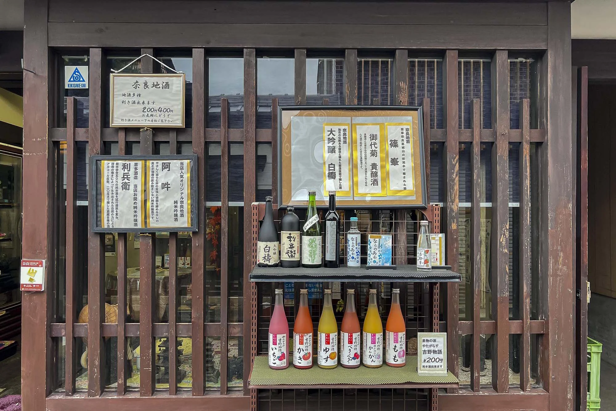 Display of various bottles of sake and alcoholic beverages on a small shelf outside a Japanese shop, with framed Japanese menus and signs on a dark wooden grid background.