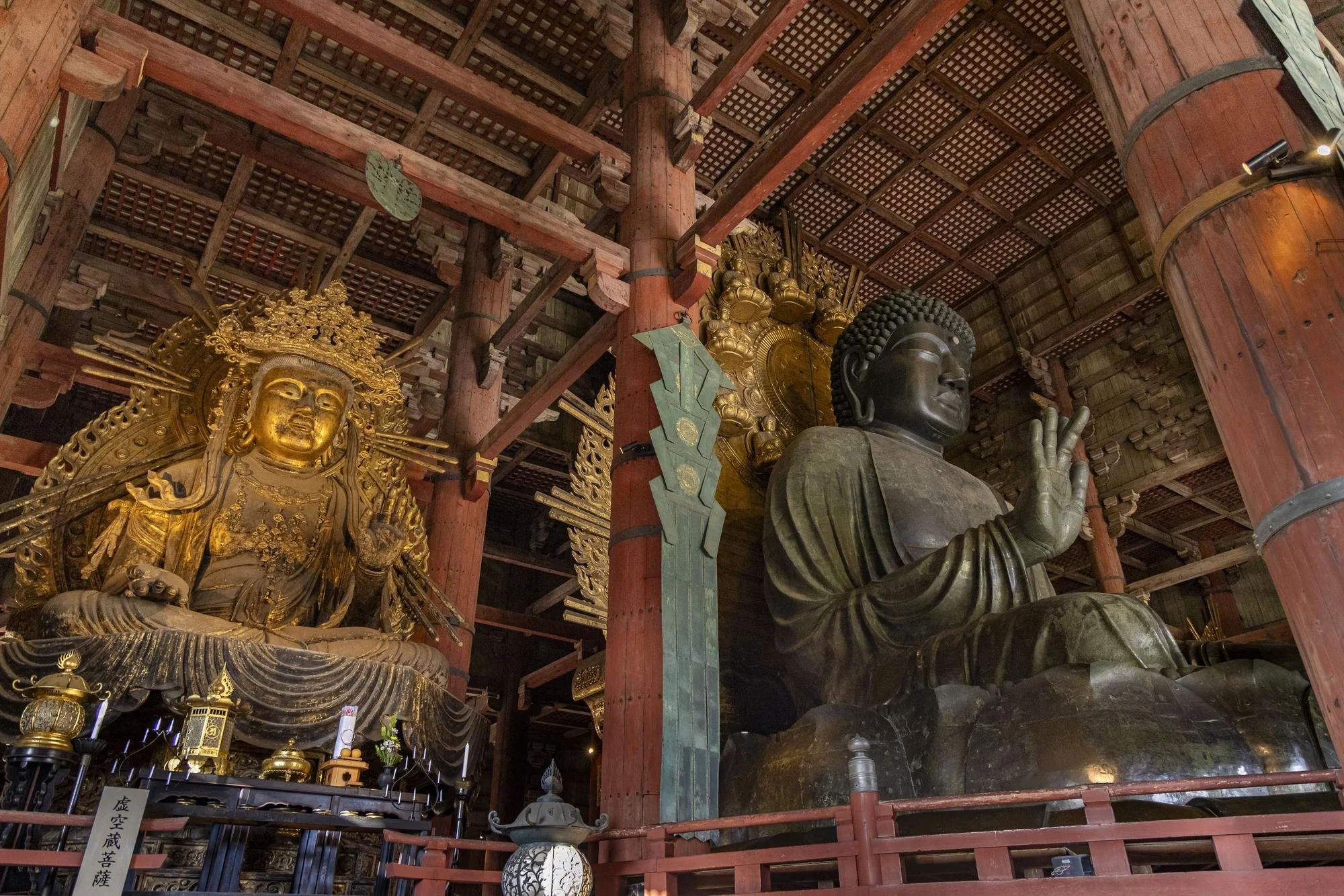 Inside a temple with large gold and bronze statues of Buddhist figures, including a seated golden deity on the left and a large bronze Buddha on the right, under a wooden ceiling with intricate beams.