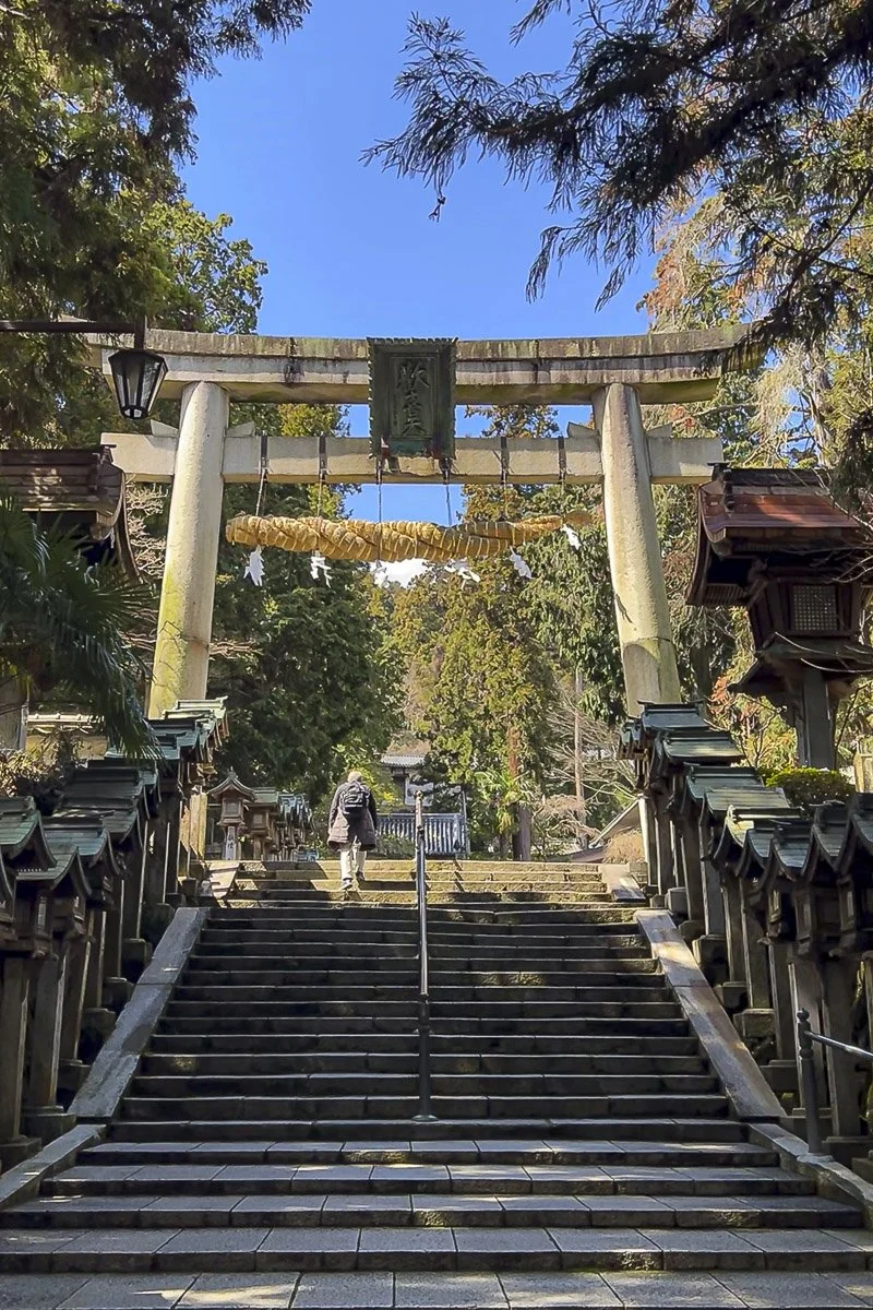 Stone steps leading up to a Shinto shrine entrance with a large torii gate at the top, surrounded by trees and traditional lanterns on the sides.