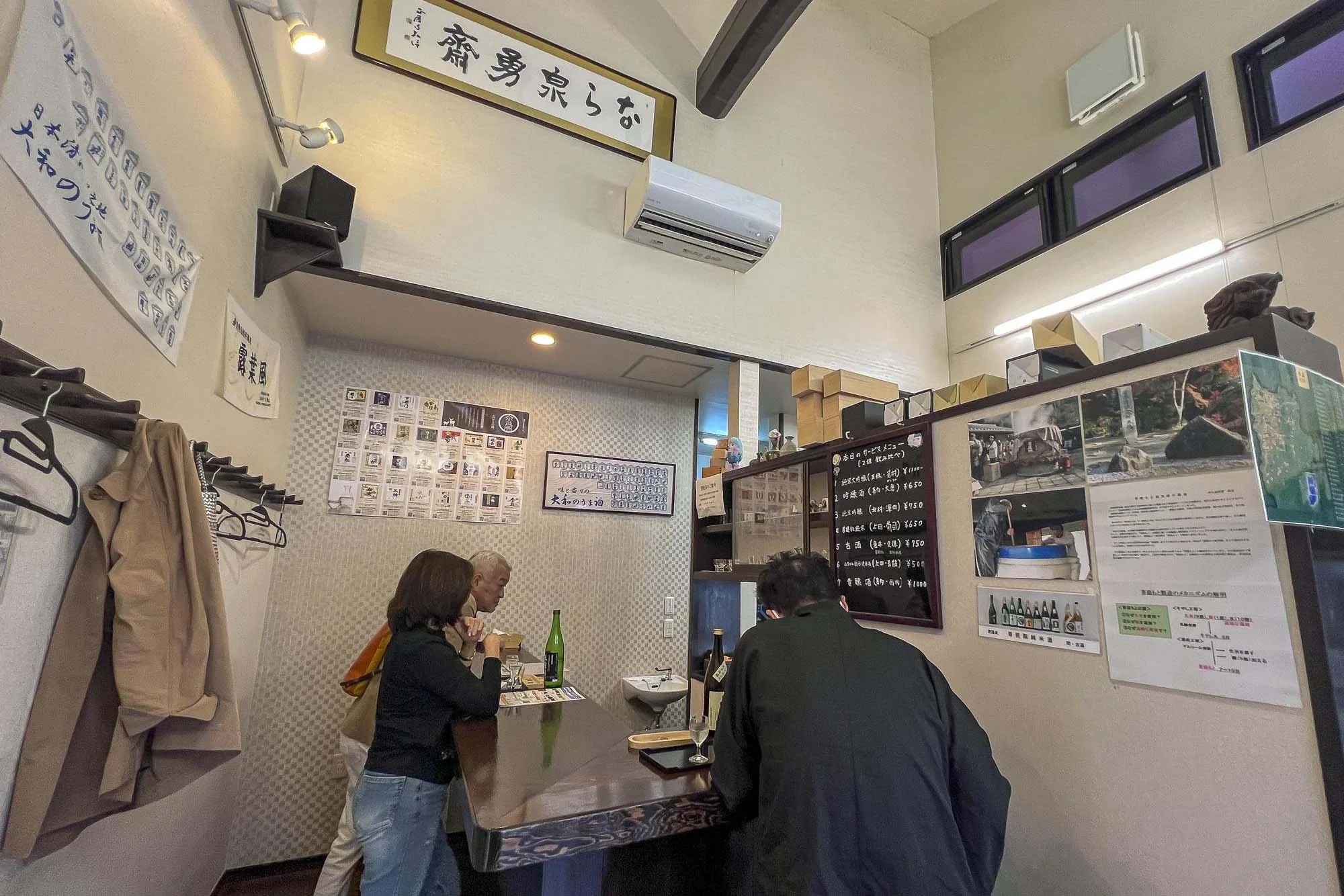 Interior of a small Japanese restaurant with three customers seated at a counter, menus on the wall, and traditional decorations.