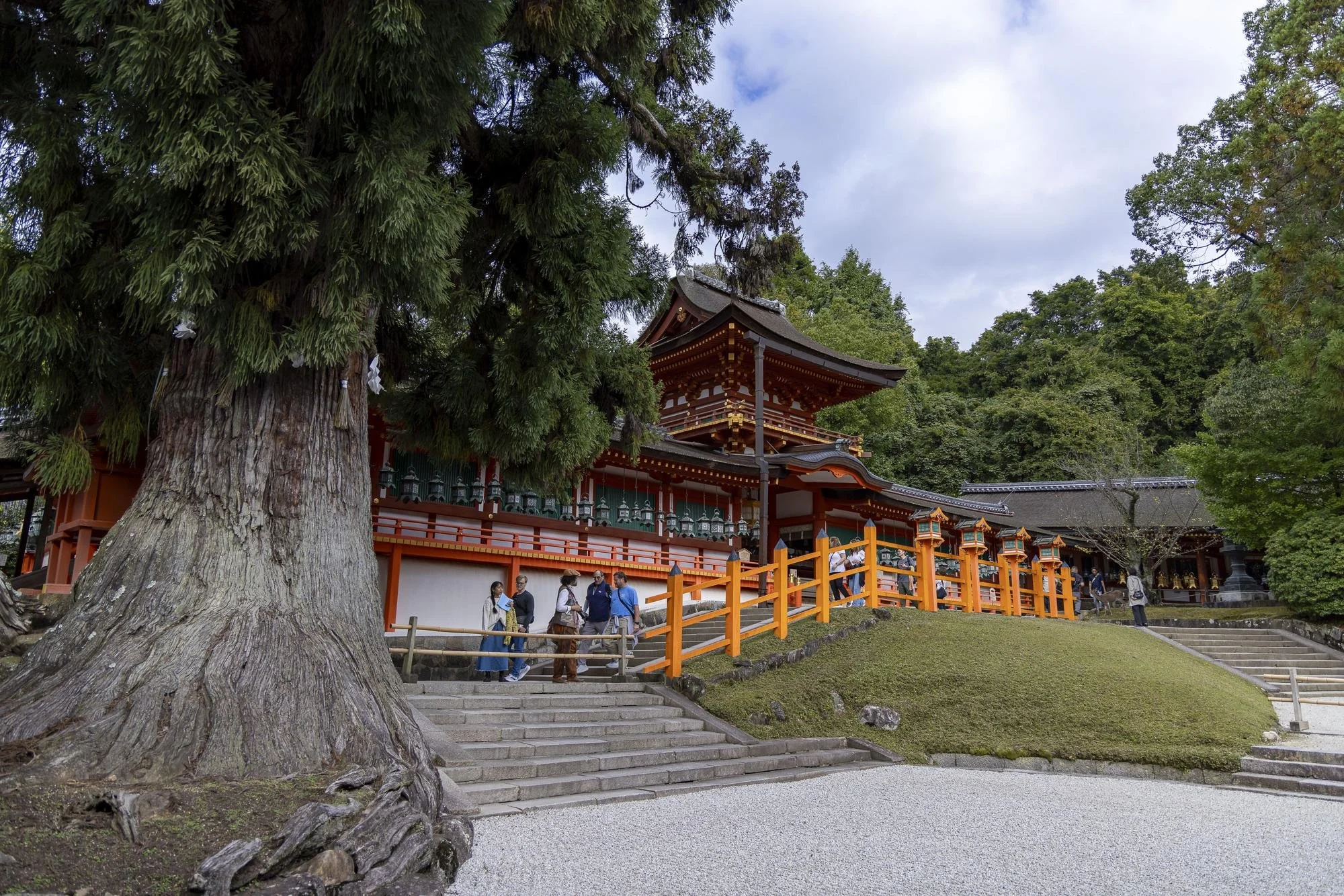 A traditional Japanese shrine with a multi-tiered roof, painted in red and black, surrounded by lush green trees. A staircase leads up to the shrine, and a group of people is walking near the entrance. The scene is set on a cloudy day.