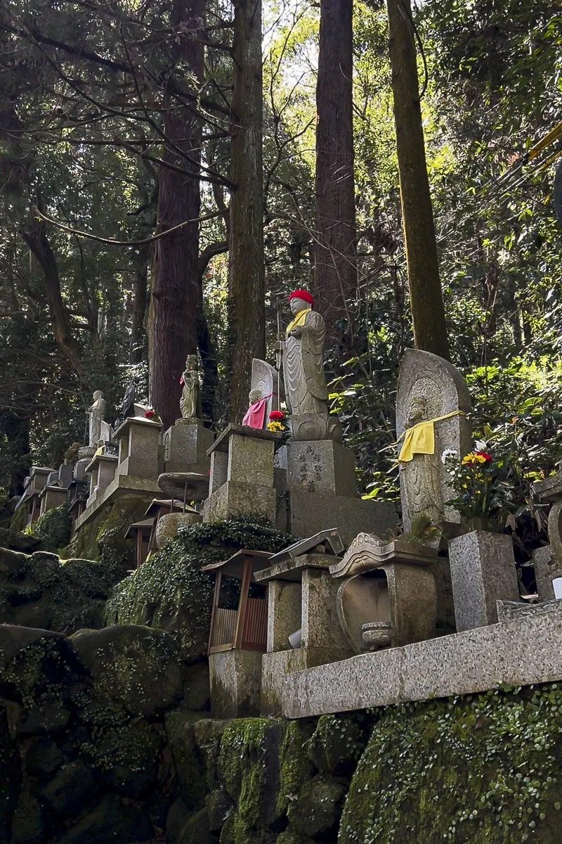 Multiple stone Buddhist statues and memorials with flowers and cloth offerings in a wooded outdoor setting.