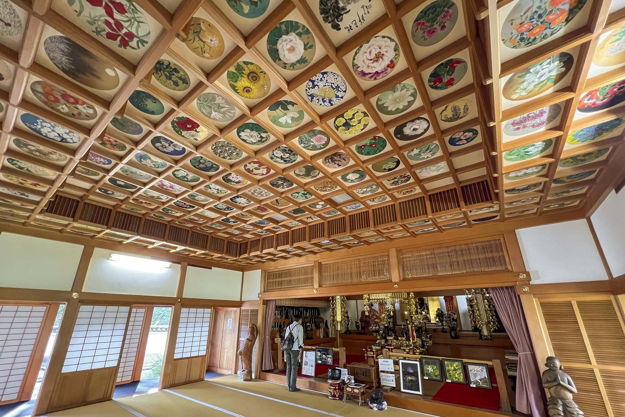 Interior of a traditional Japanese room with a colorful, decorated ceiling featuring circular patterns of floral and nature motifs, wooden sliding doors, and an altar with religious artifacts and statues.
