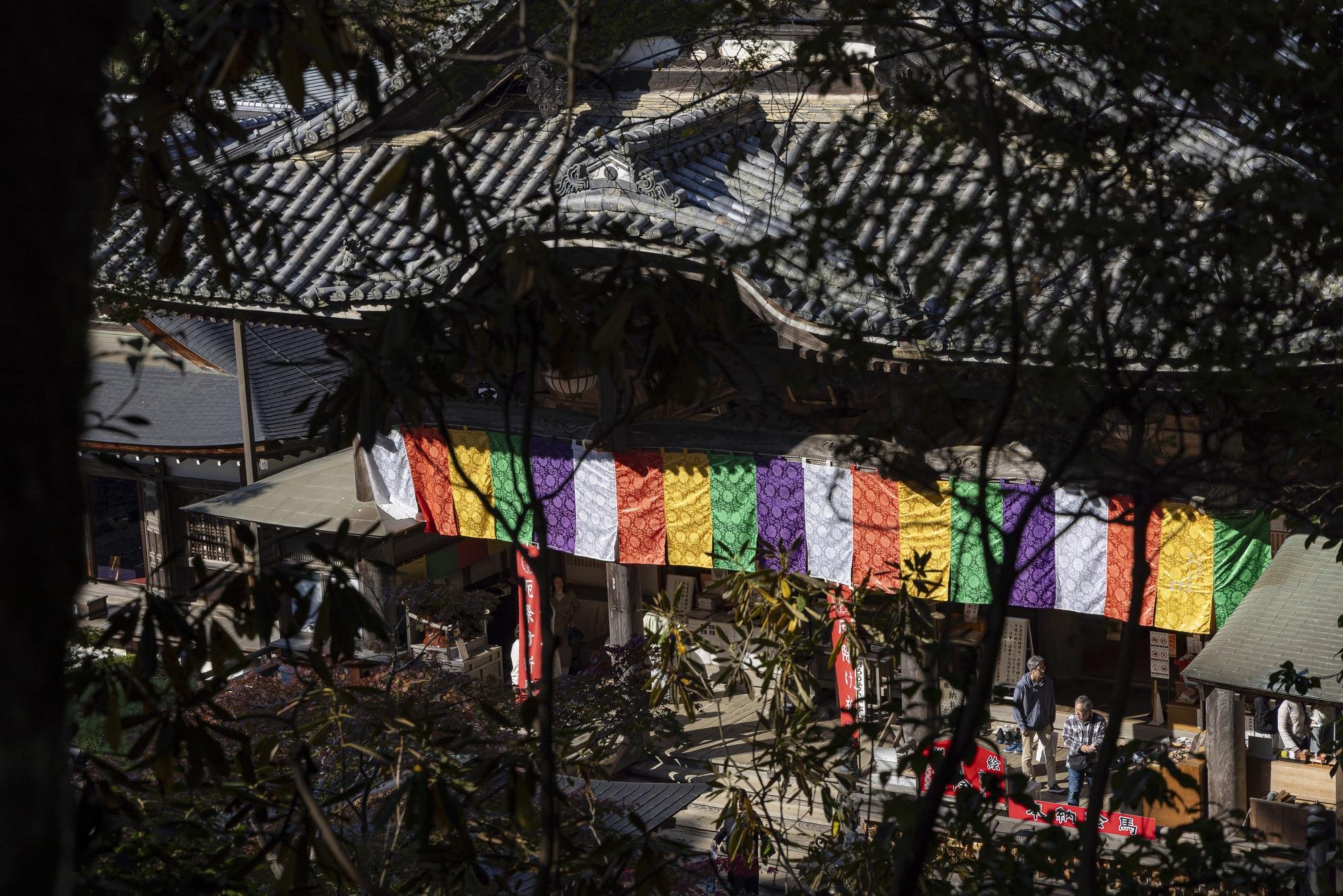 A traditional Japanese temple decorated with colorful paper banners, viewed through tree branches.
