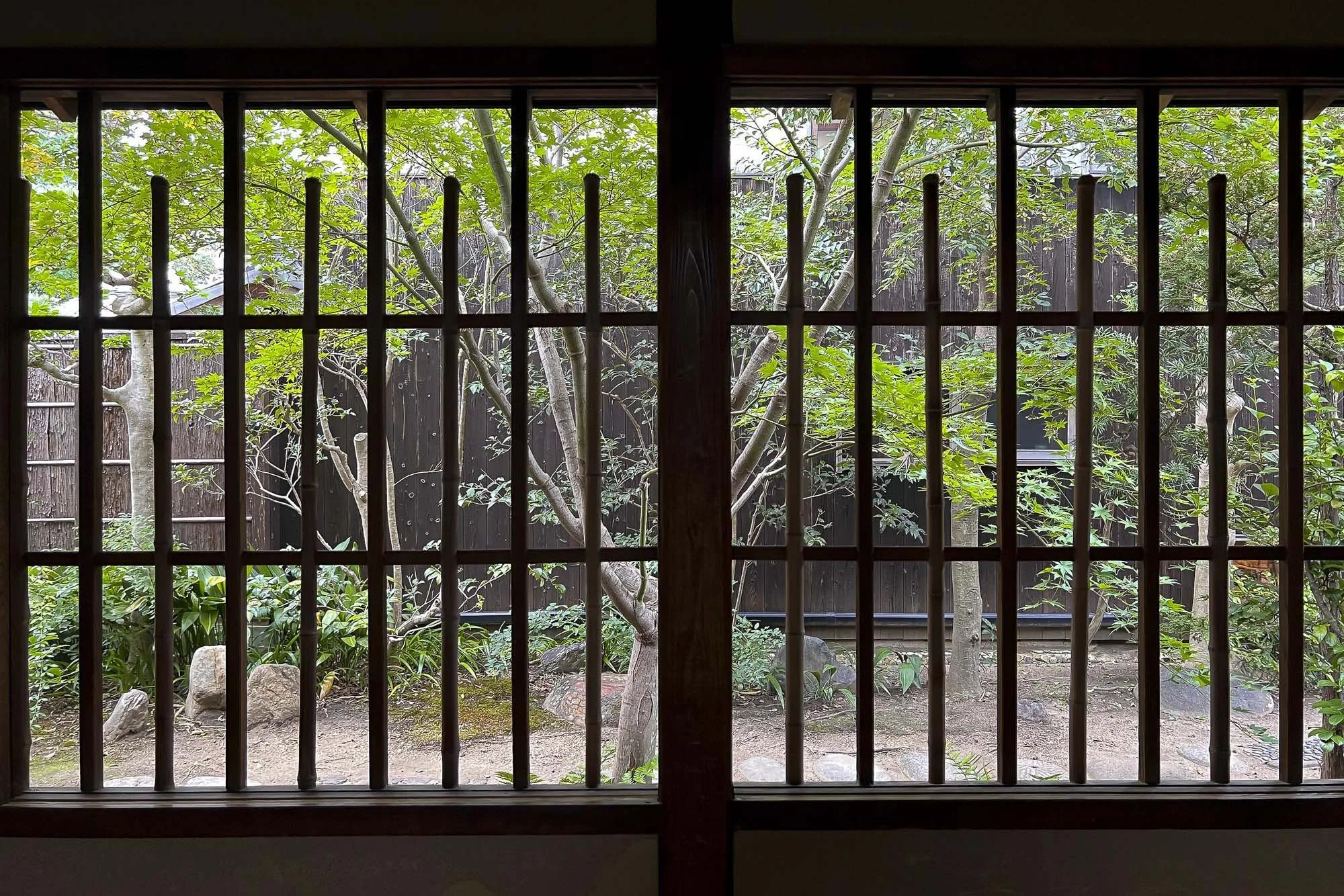 View of a garden through traditional Japanese wooden window with grid pattern, featuring green trees, plants, and rocks.