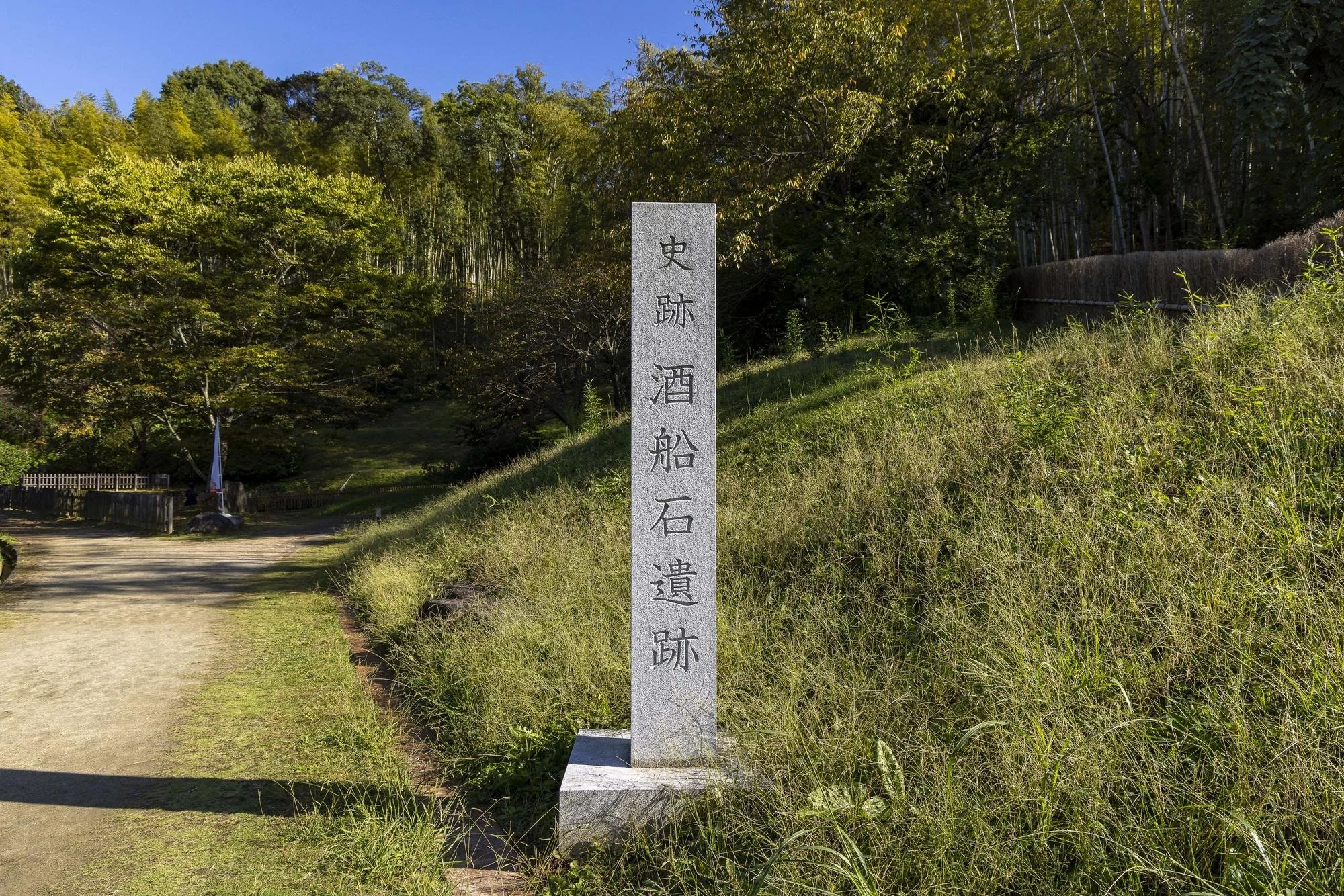 A stone monument with Japanese inscriptions standing on grassy terrain at a historical site, with trees and a dirt path in the background.