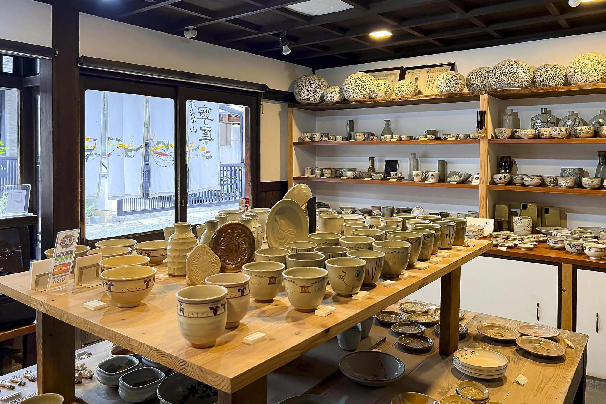 Interior view of a pottery shop with shelves filled with handcrafted ceramic bowls, plates, and vases. A wooden table displays numerous ceramic cups and bowls, with more ceramics arranged on the shelves and the table.