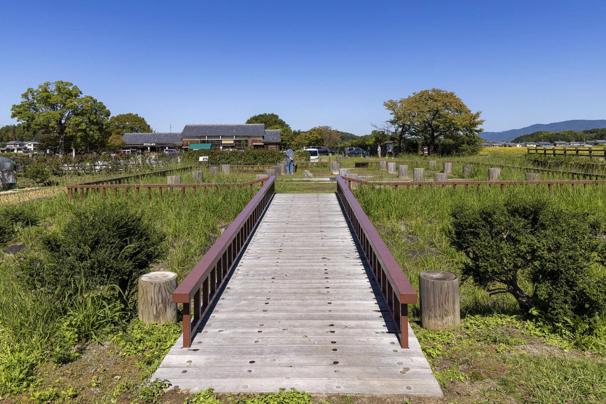 A wooden footbridge extending over a grassy area, leading to a park with trees and a few people in the distance under a clear blue sky.