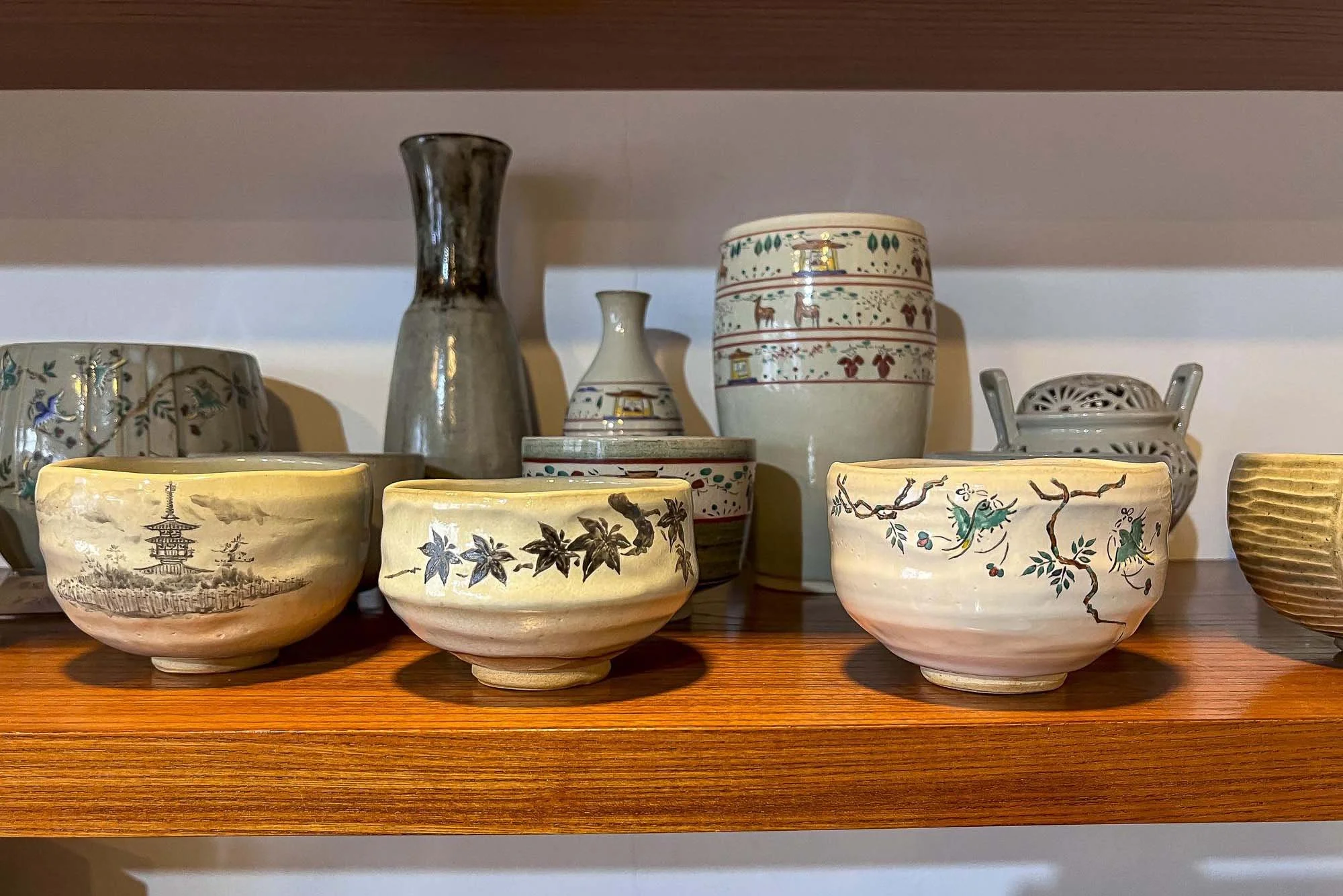 A wooden shelf holding various ceramic bowls and vases with traditional Japanese designs, a tall black and silver vase, and a smaller white vase in the background.