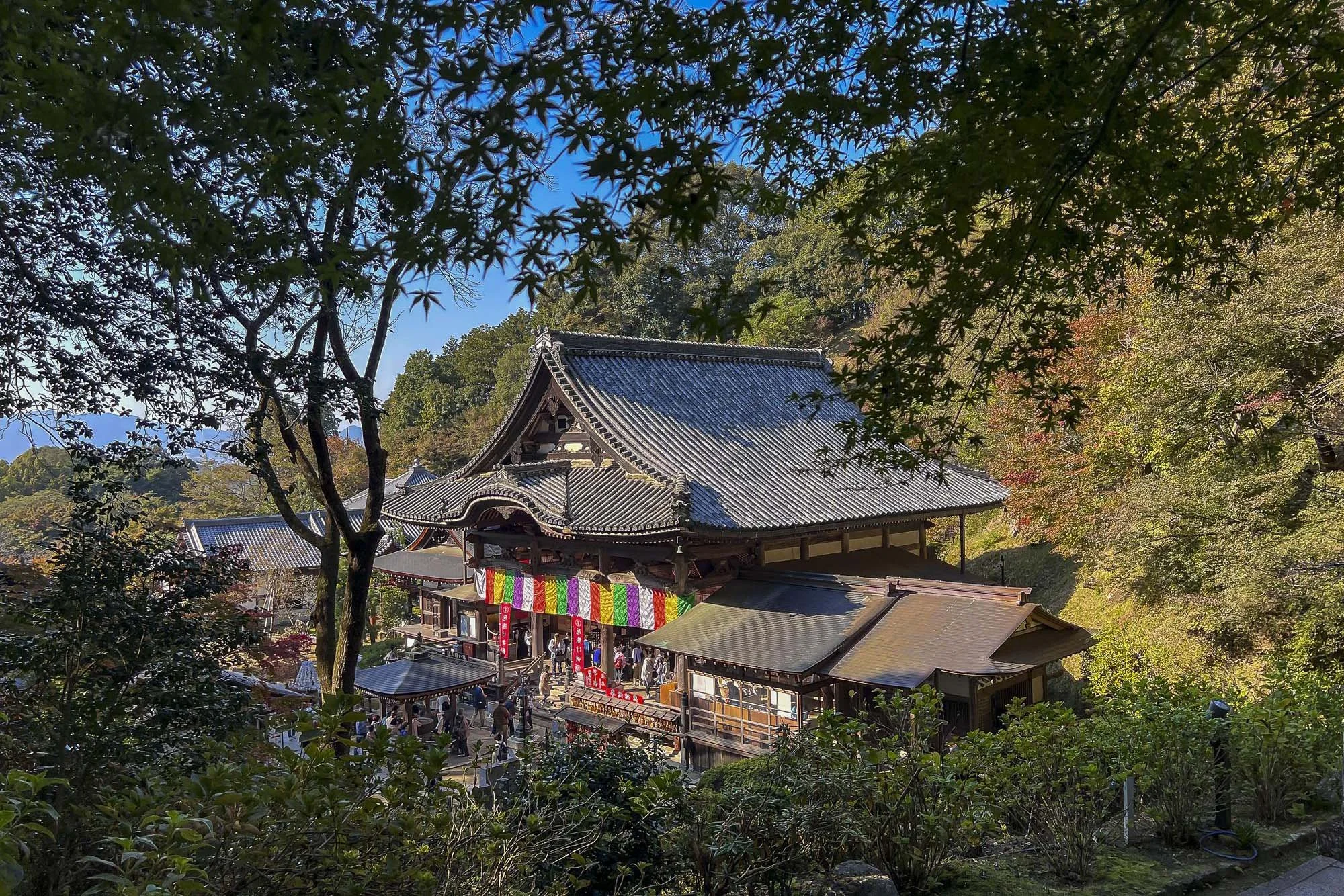 A traditional Japanese wooden temple with colorful banners, surrounded by trees and visitors, under a clear blue sky.
