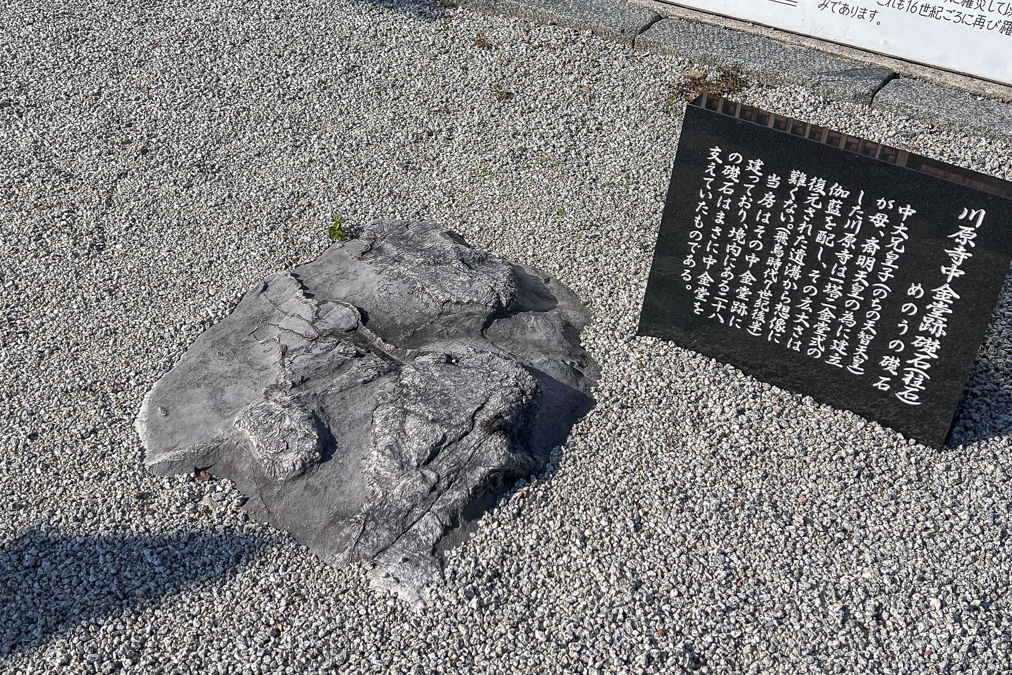 A large, dark gray stone with carved symbols or inscriptions on its surface, placed on white gravel. Next to it is a black informational plaque with Japanese text explaining the historical significance of the site.