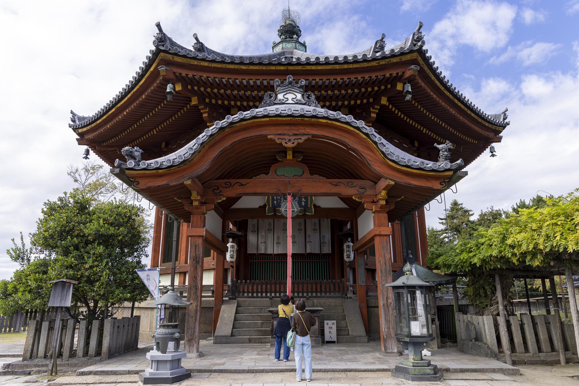 A traditional Japanese shrine with a curved wooden roof and steps leading up to the entrance, two visitors are standing in front of it, and there are lanterns and trees around.
