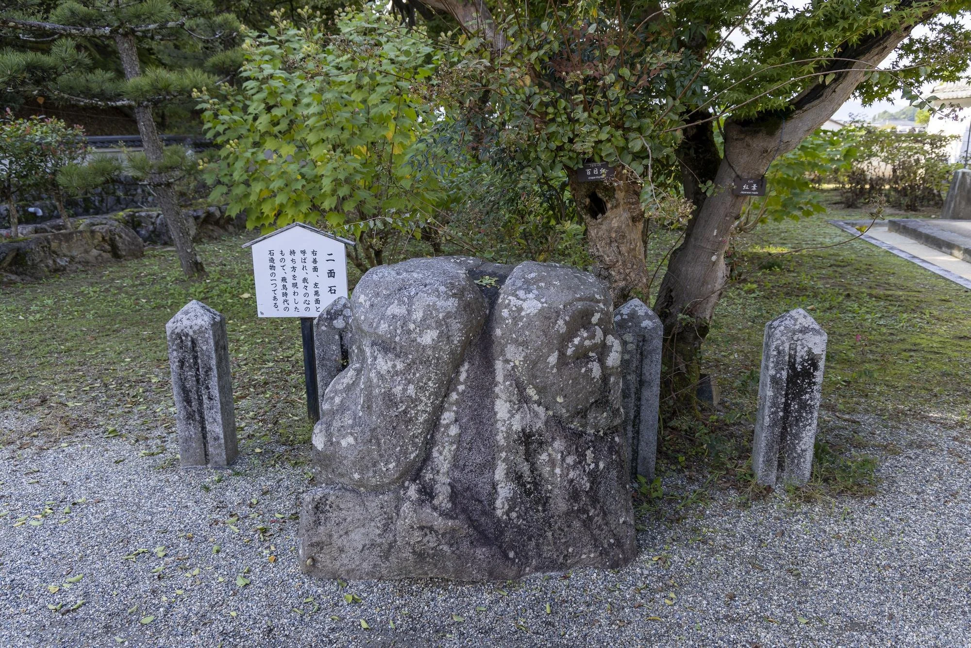 A large landscape stone with an abstract face carving, surrounded by four smaller posts, with a white informational sign in Japanese nearby, set in a garden with trees and grass.