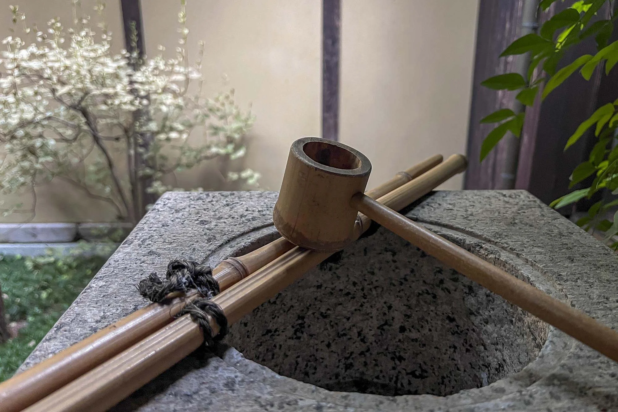 A traditional Japanese stone water basin with a bamboo dipper and a wooden mallet resting on it, set outdoors with plants in the background.