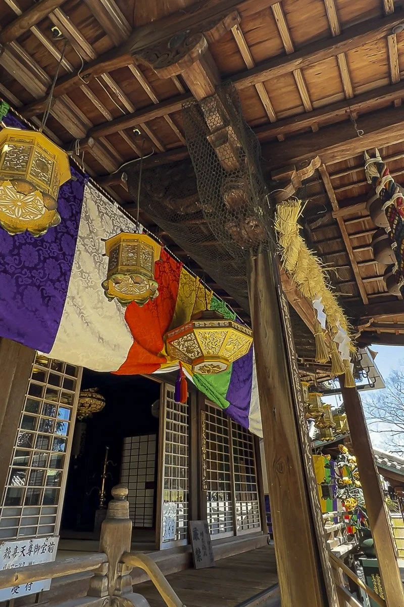 Colorful traditional lanterns hanging under a wooden temple roof with purple, white, red, green, and yellow fabrics.