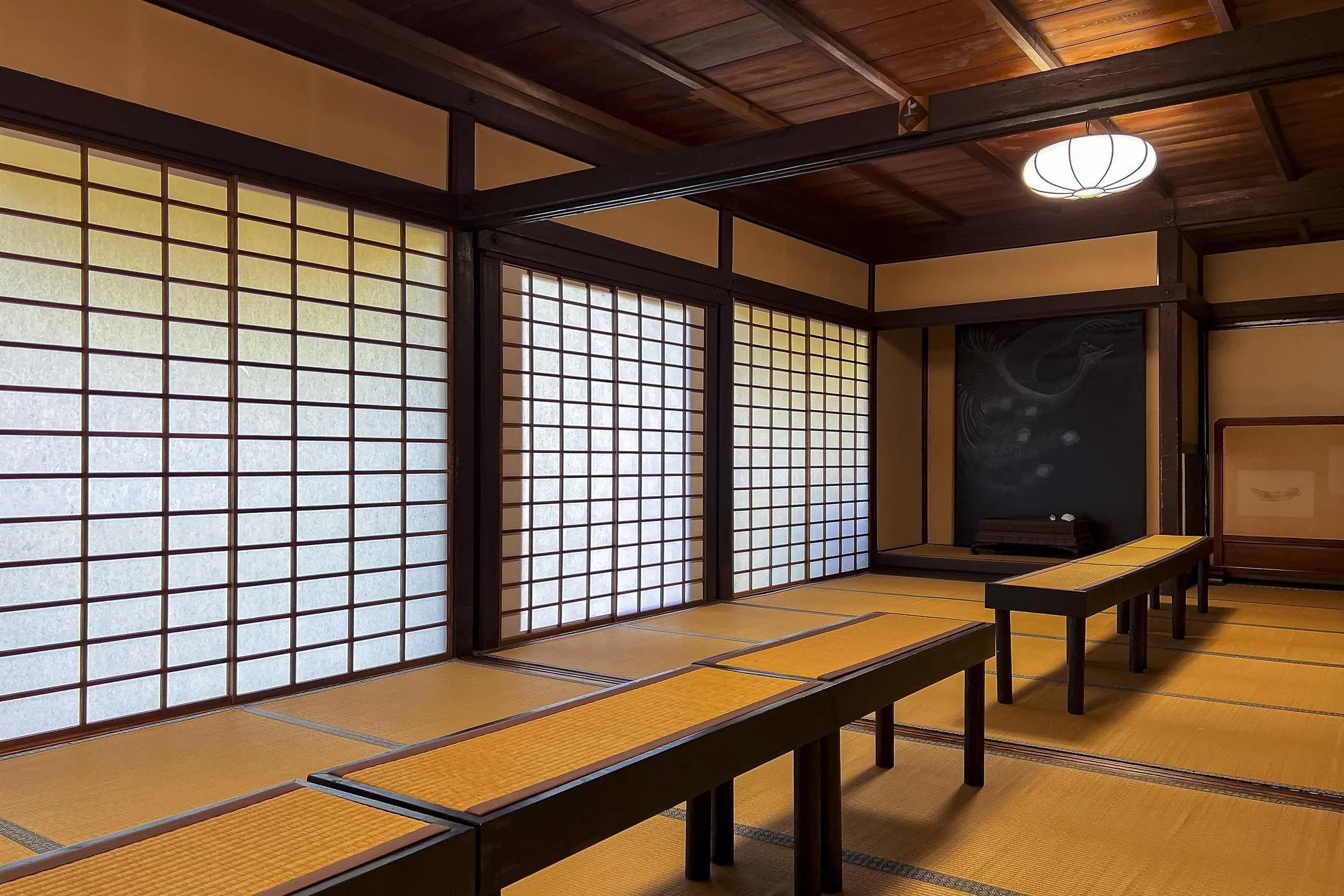 A traditional Japanese room with tatami mat flooring, shoji sliding doors, a dark wooden ceiling, and a black mural of a dragon or phoenix on the back wall.