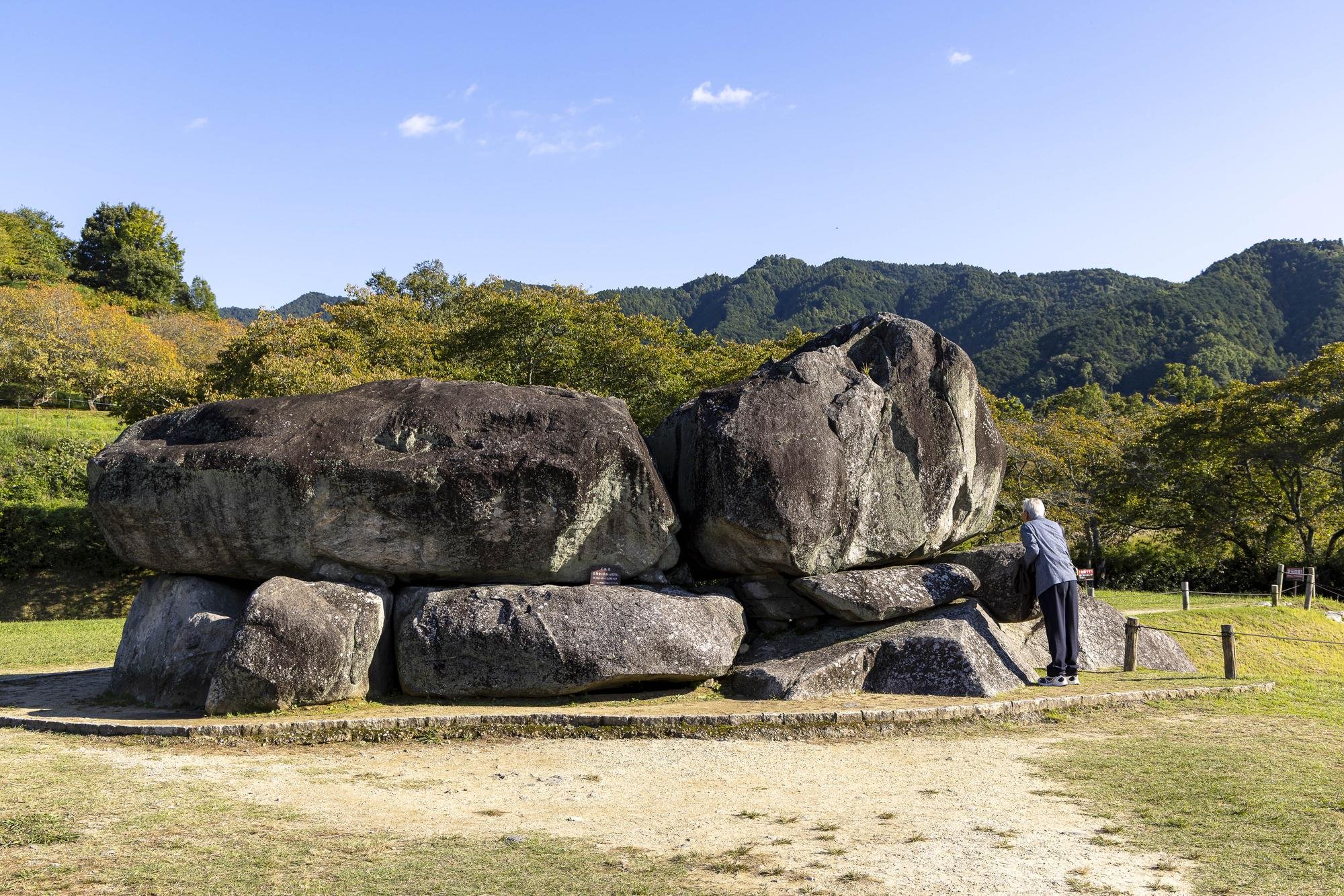 A woman stands near large boulders on a grassy area with trees and mountain hills in the background under a clear blue sky.