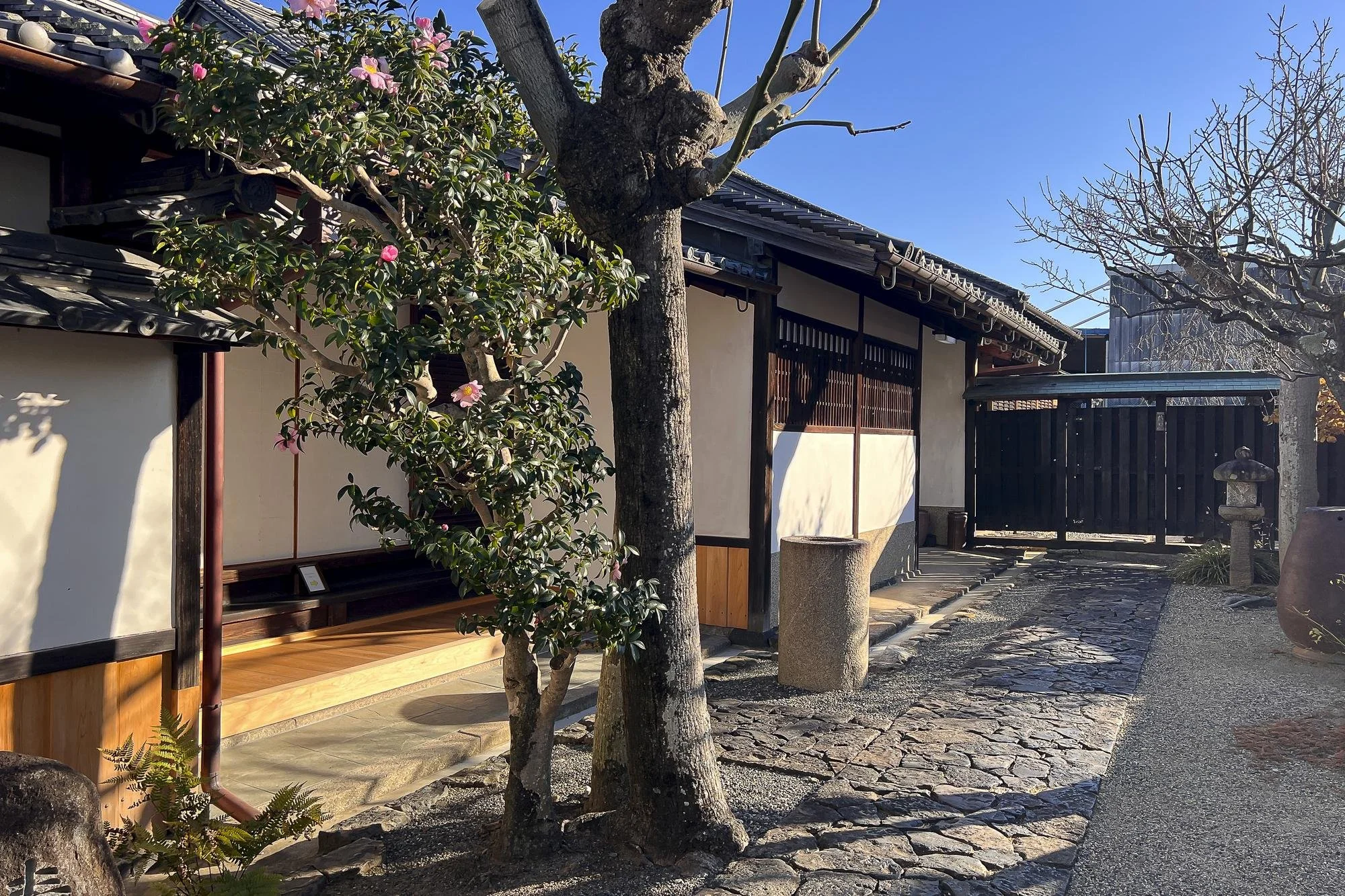 A traditional Japanese courtyard with a stone pathway, a wooden building with sliding doors, and trees including one with pink flowers, under a clear blue sky.