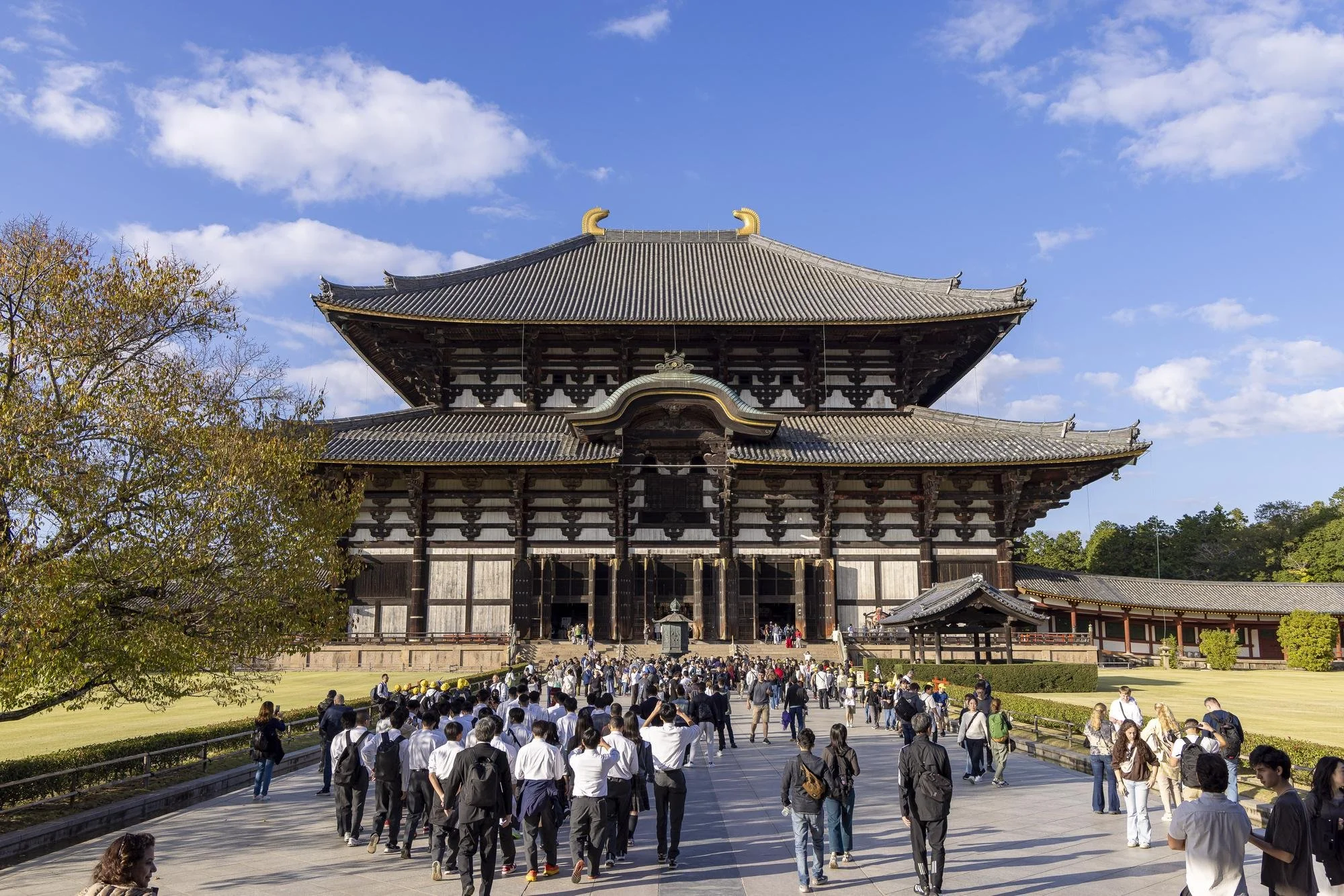A large traditional Japanese temple with many visitors walking towards it on a sunny day. The temple is multi-storied with a curved roof and intricate wooden details. There are trees and a clear blue sky in the background.