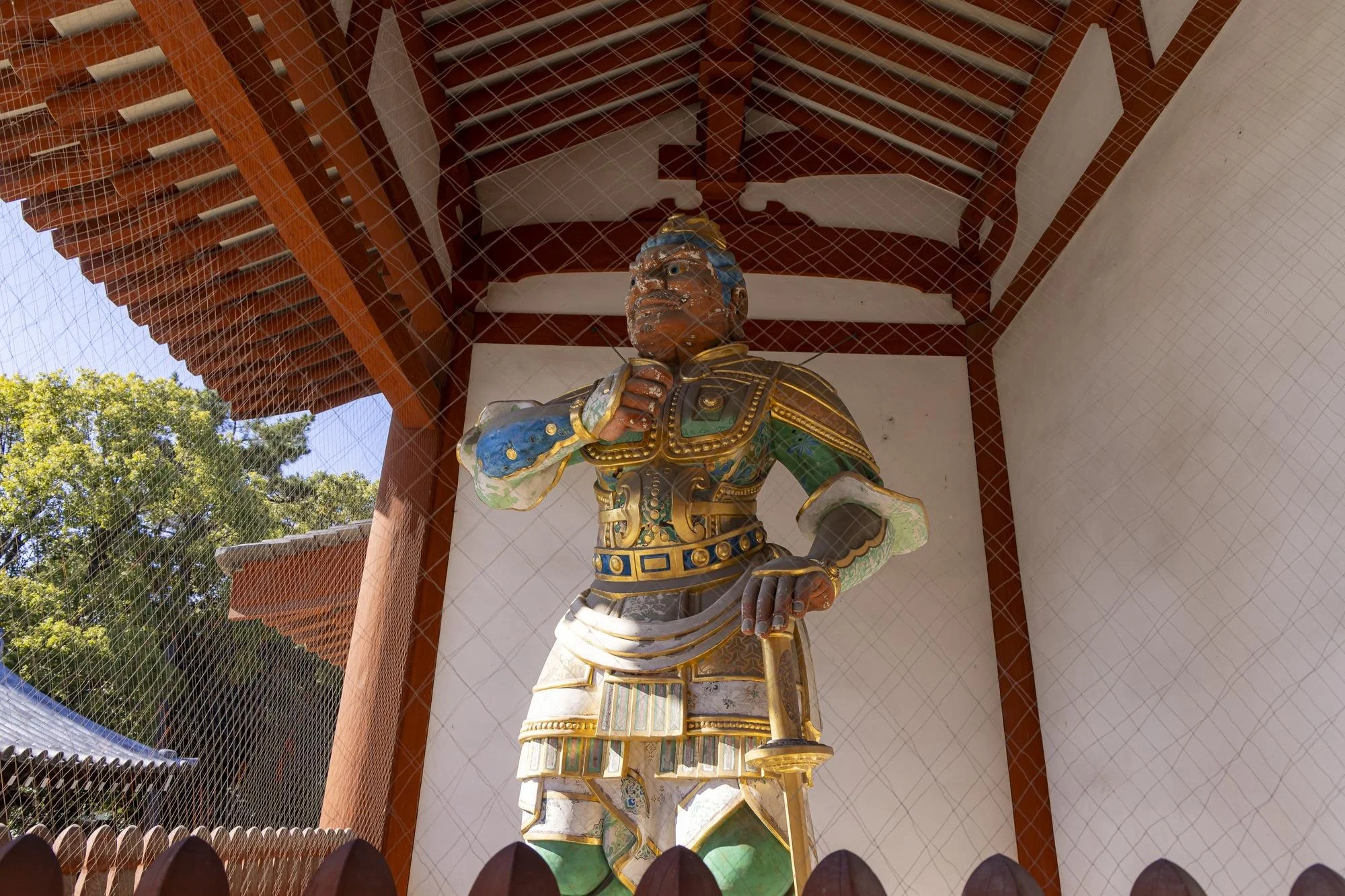 A large, colorful statue of a warrior in traditional armor, holding a sword and standing on a platform inside a wooden structure with a roof, surrounded by trees.