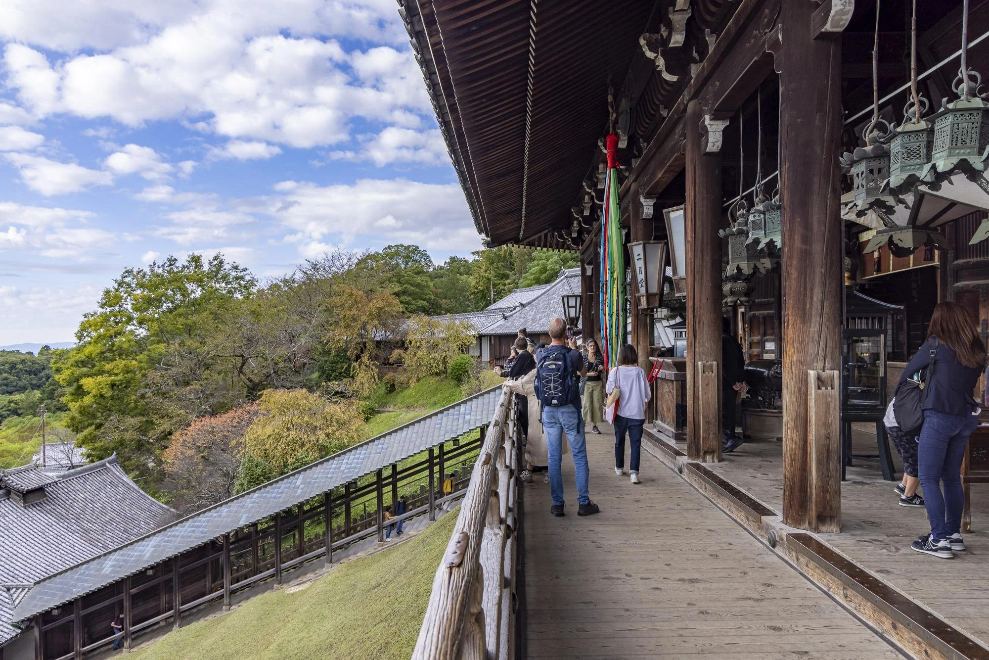 People sightseeing on the wooden balcony of a traditional Japanese temple or shrine, with trees and rooftops in the background under a partly cloudy sky.