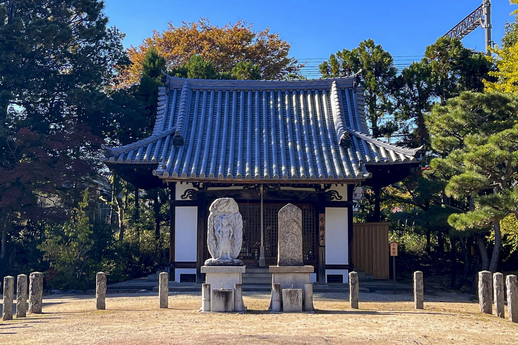 A traditional Japanese shrine with a blue-tiled roof, surrounded by trees with autumn foliage, and two stone statues in front.