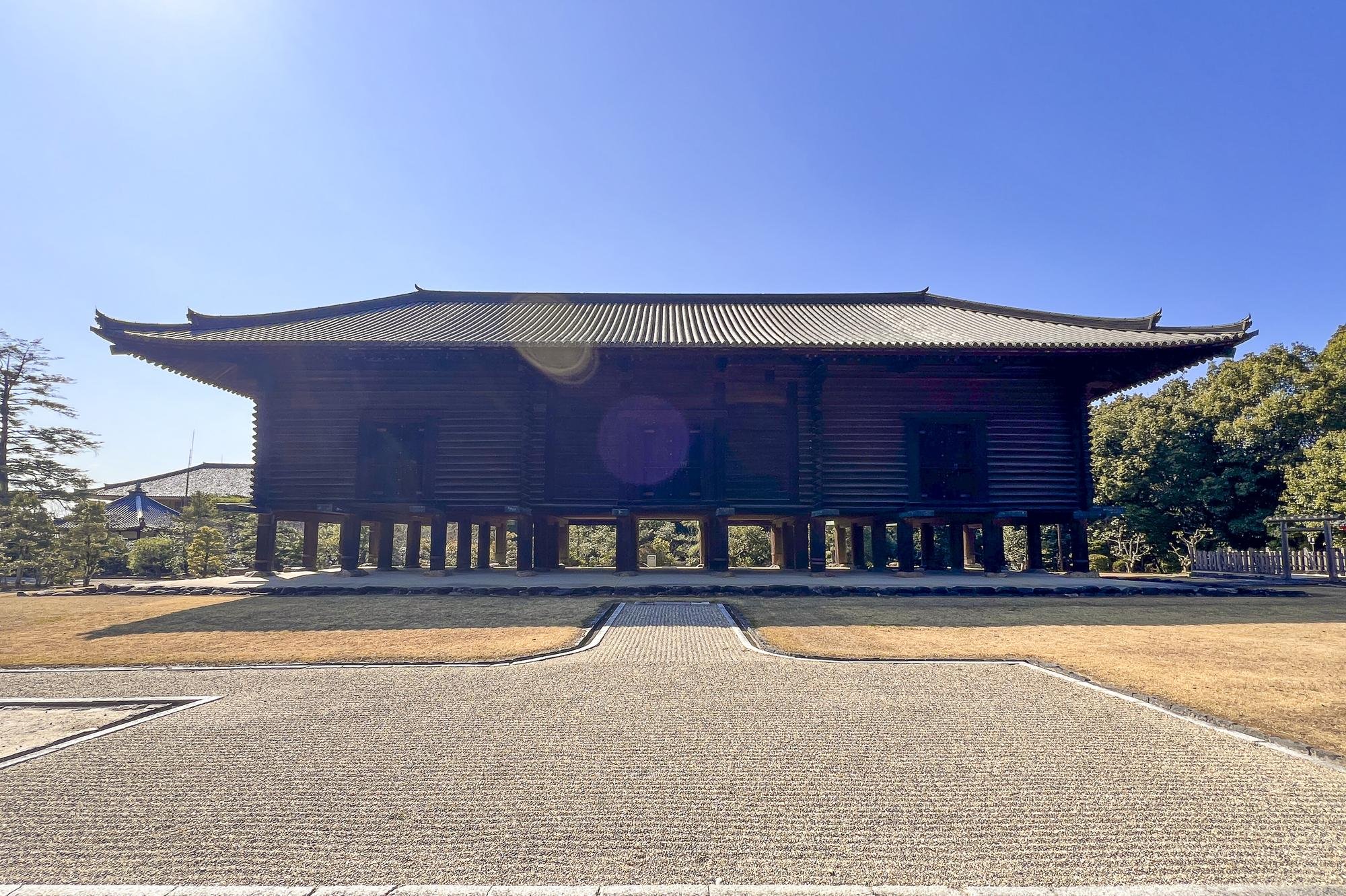 Traditional Japanese wooden building on stilts with a tiled, curved roof, surrounded by greenery and a clear sky.